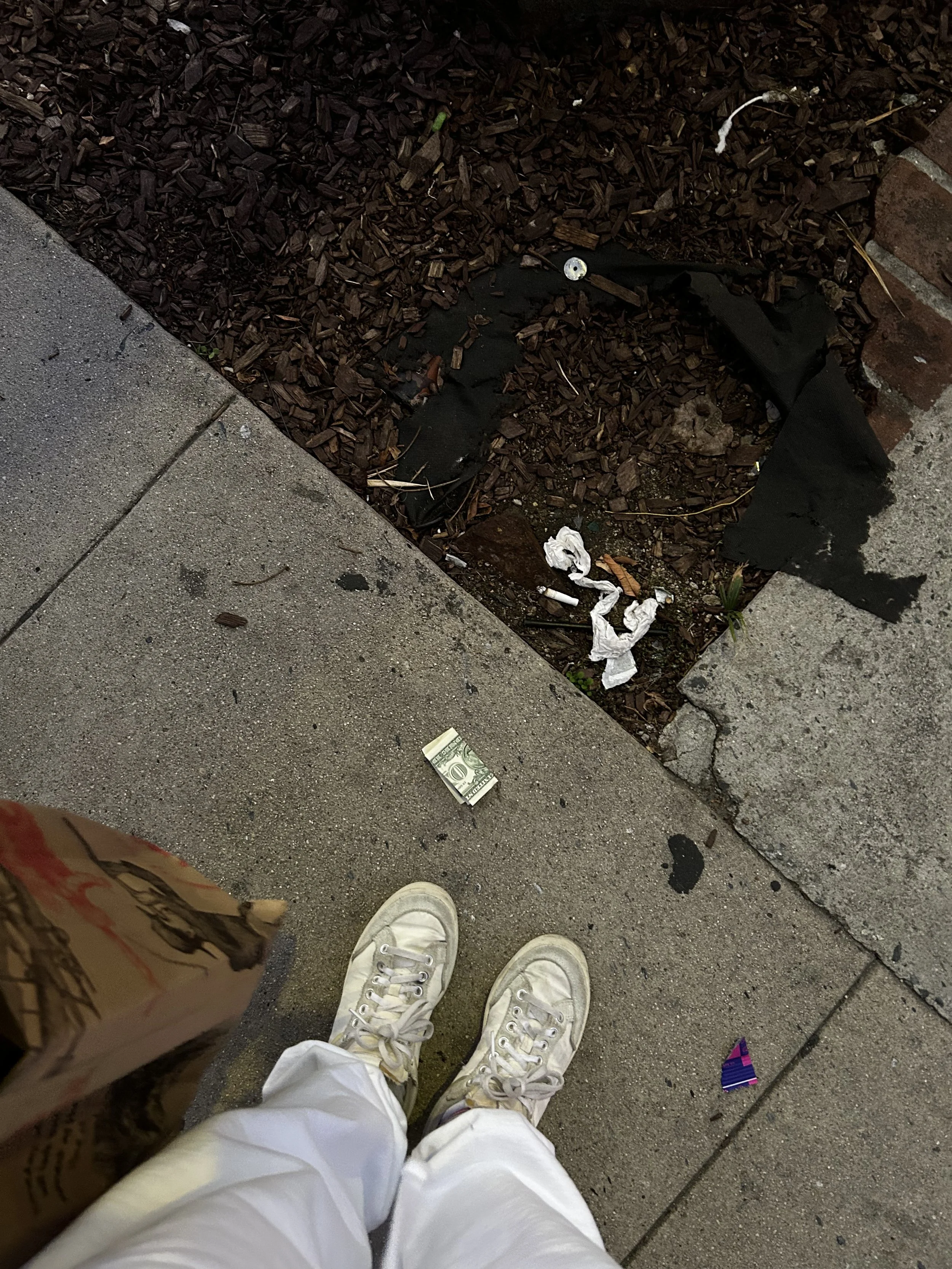 A street-level shot of worn white sneakers beside a crumpled bill on the sidewalk, framed by a paper grocery bag — a raw, unfiltered Shy Rockstar moment.