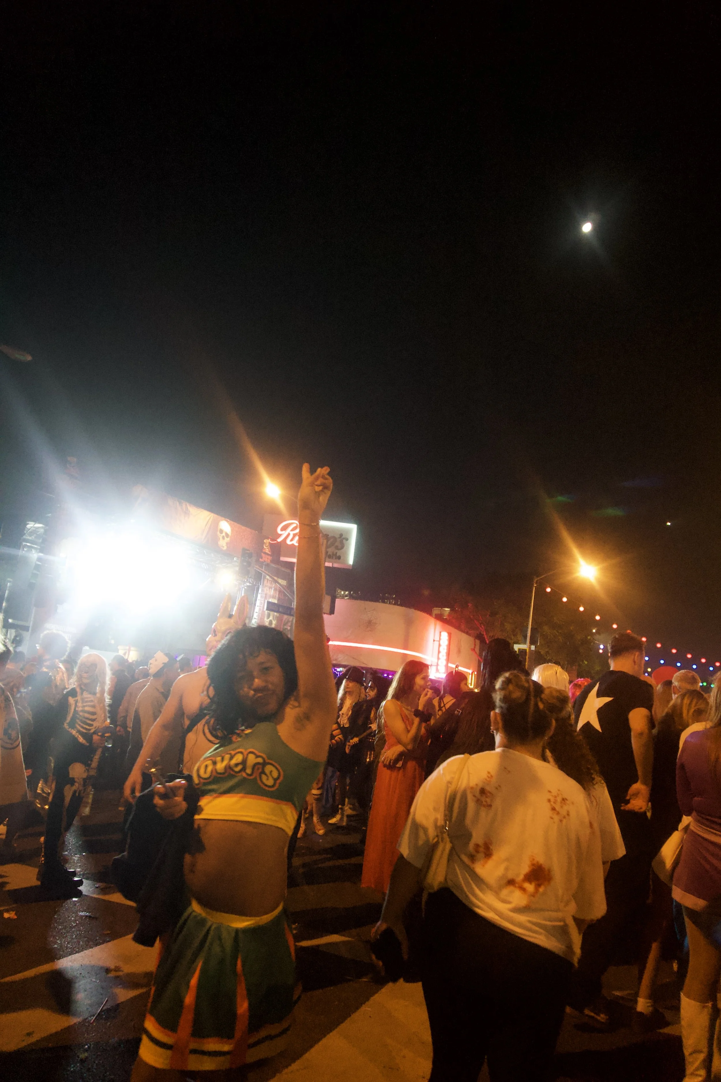 Night street scene of a lively crowd under city lights, with a costumed man raising his hand toward the moon, capturing the spontaneous, cinematic chaos of Shy Rockstar Studios.