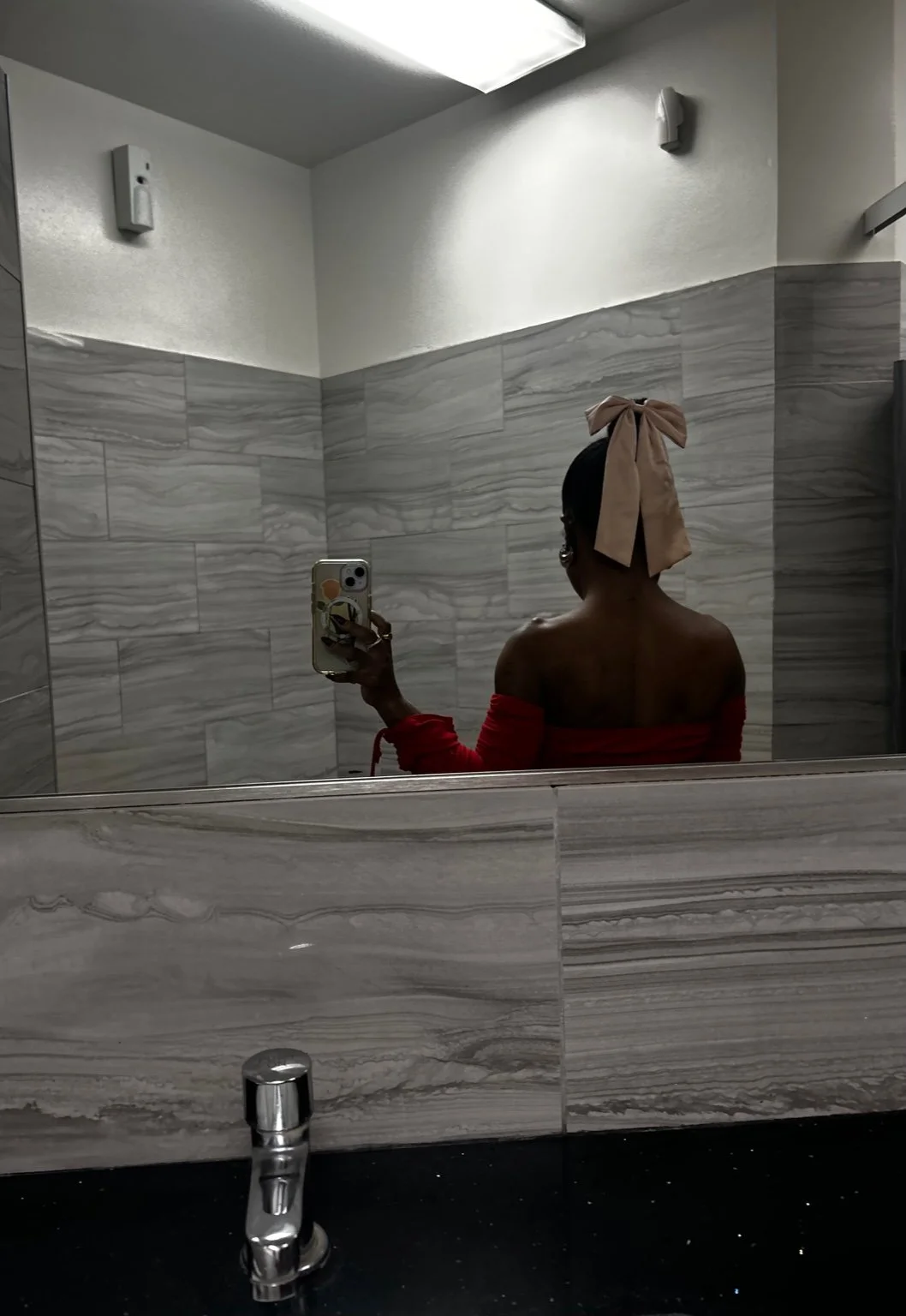 Person wearing an off-the-shoulder red dress taking a mirror selfie in a tiled restroom, hair styled in a low bun with a large bow.