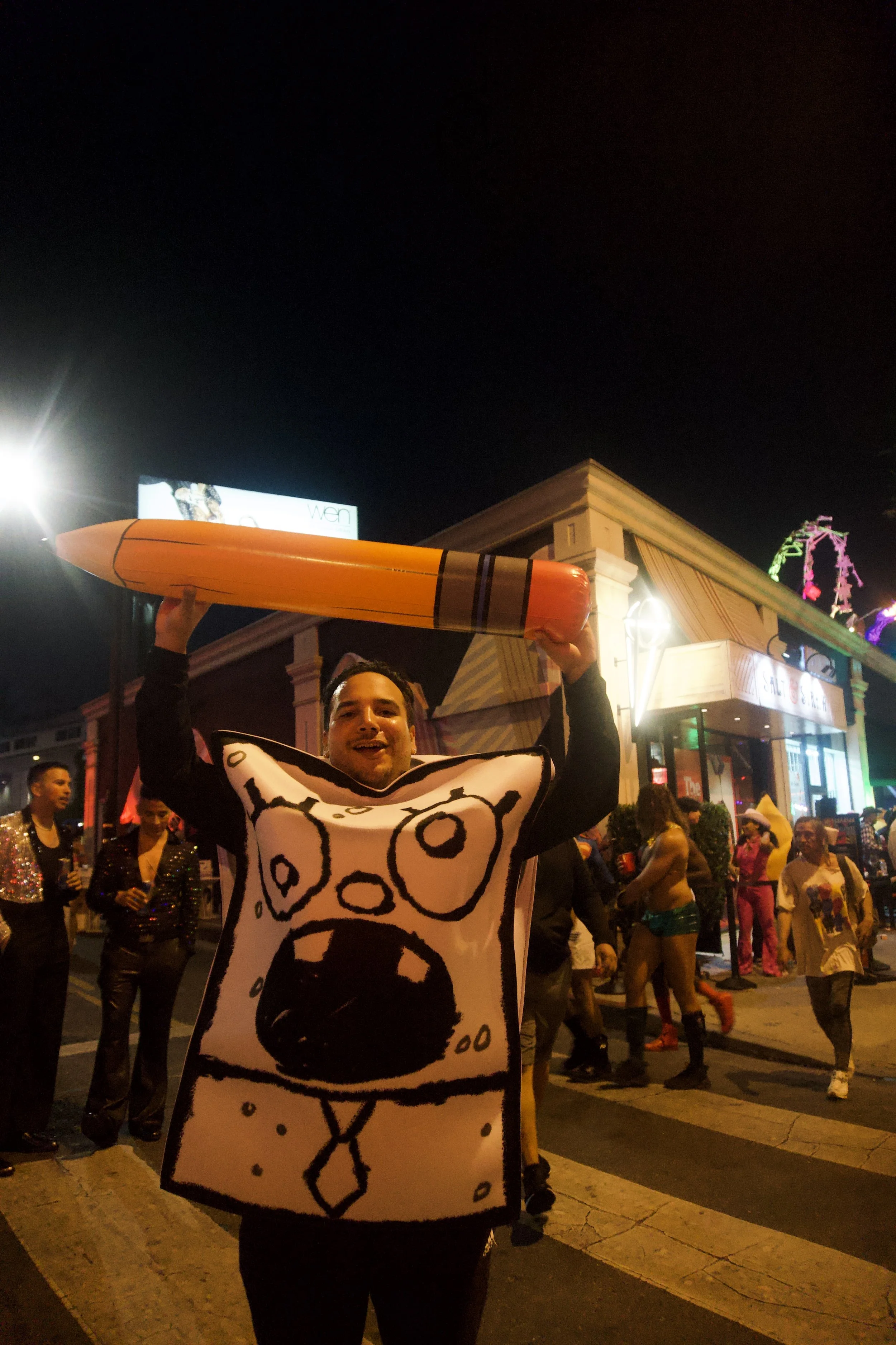 Person in a playful cartoon-style costume holding a giant inflatable crayon during a lively night street celebration, capturing the colorful, energetic vibe of Shy Rockstar Studios.