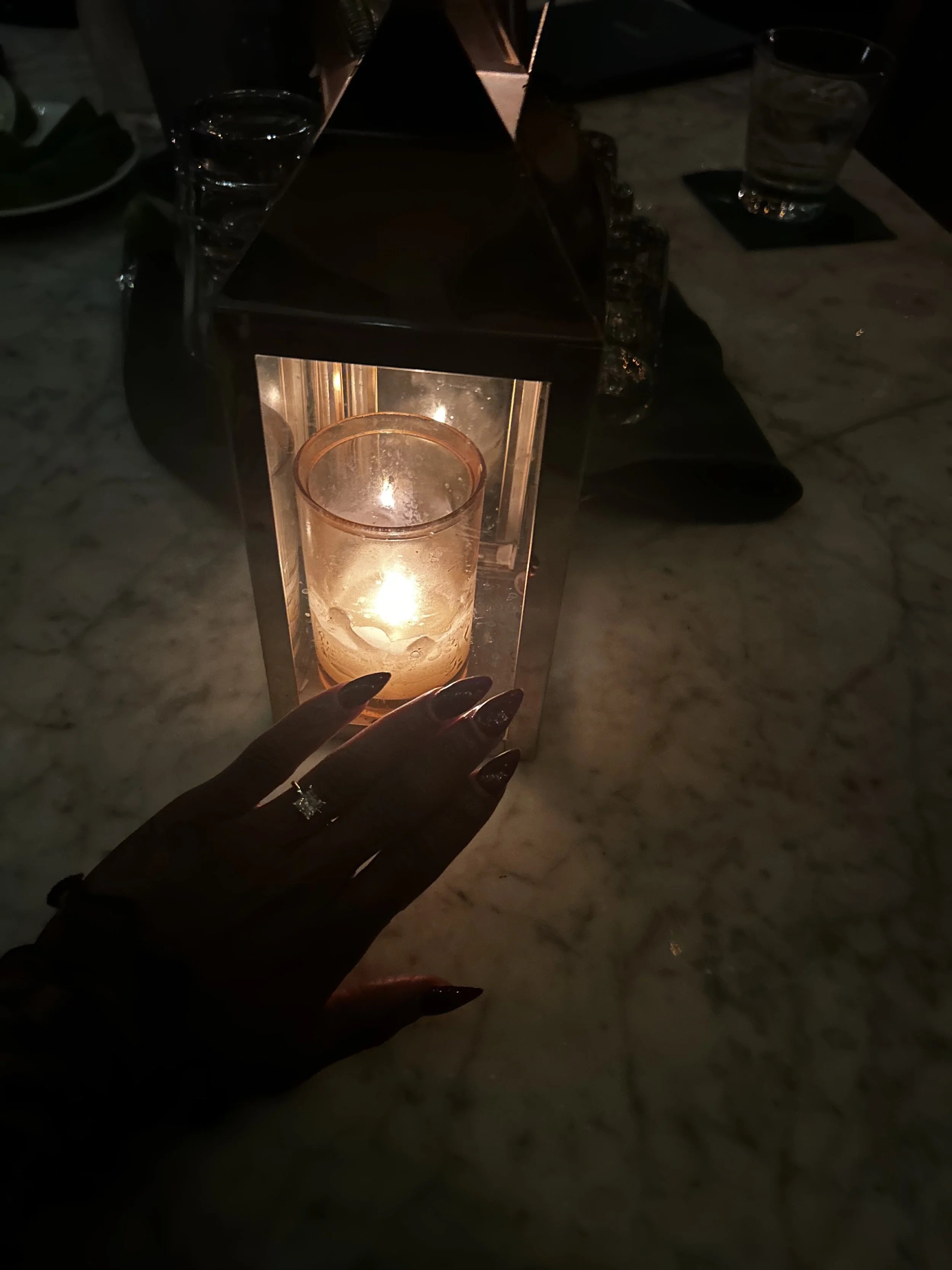 A hand with an engagement ring reaches toward a lit candle inside a lantern on a marble table.