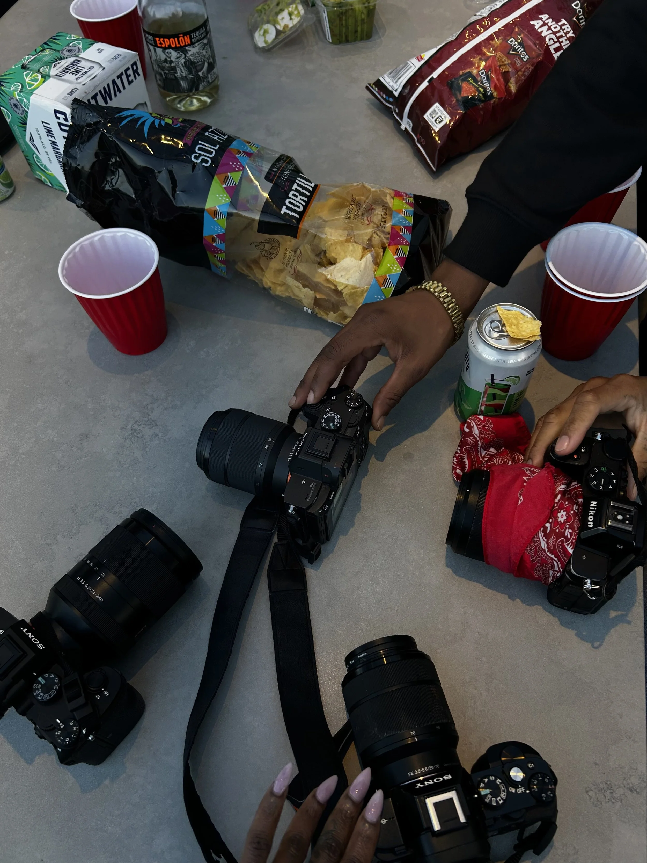 Several cameras on a table with snacks and drinks, including tortilla chips, a soda can, an alcohol shot, and red cups, with two people handling a camera.