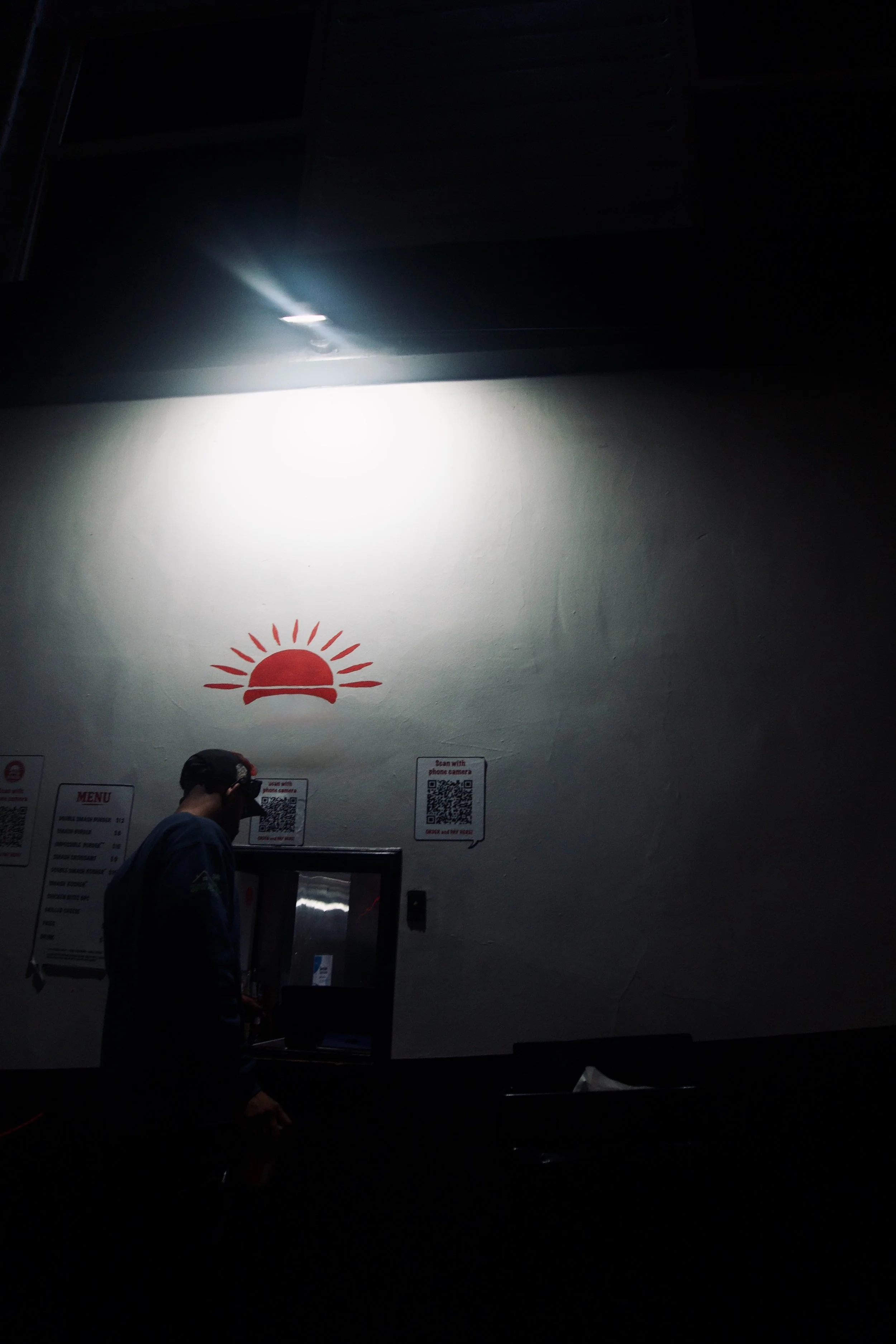 Cinematic night scene of a man walking past a bright spotlight and a red sun symbol painted on a wall, capturing the quiet, moody ambience of Shy Rockstar Studios.