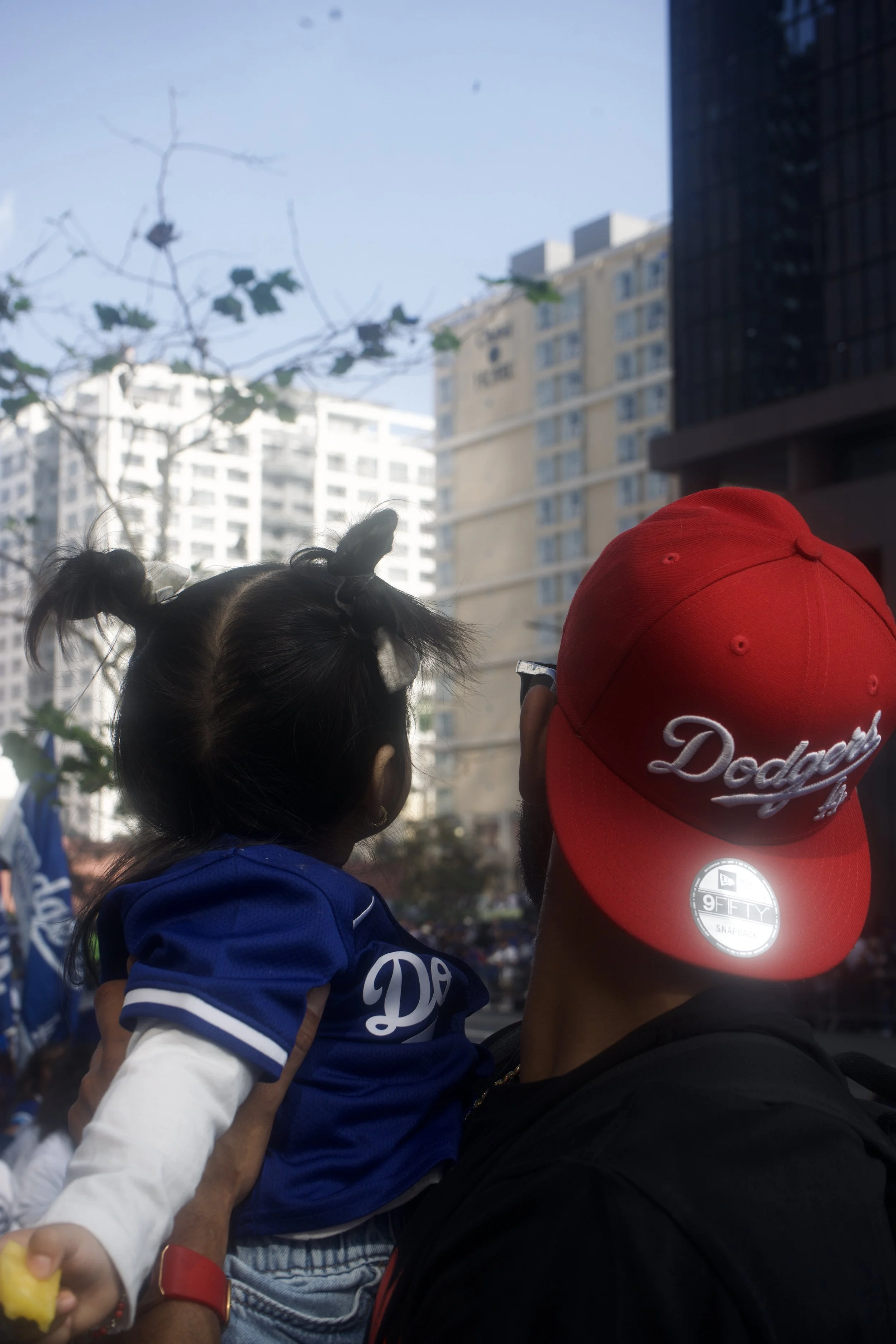 Adult holding a child in matching Dodgers gear during a daytime celebration, with city buildings in the background, capturing a warm, candid moment in true Shy Rockstar style.