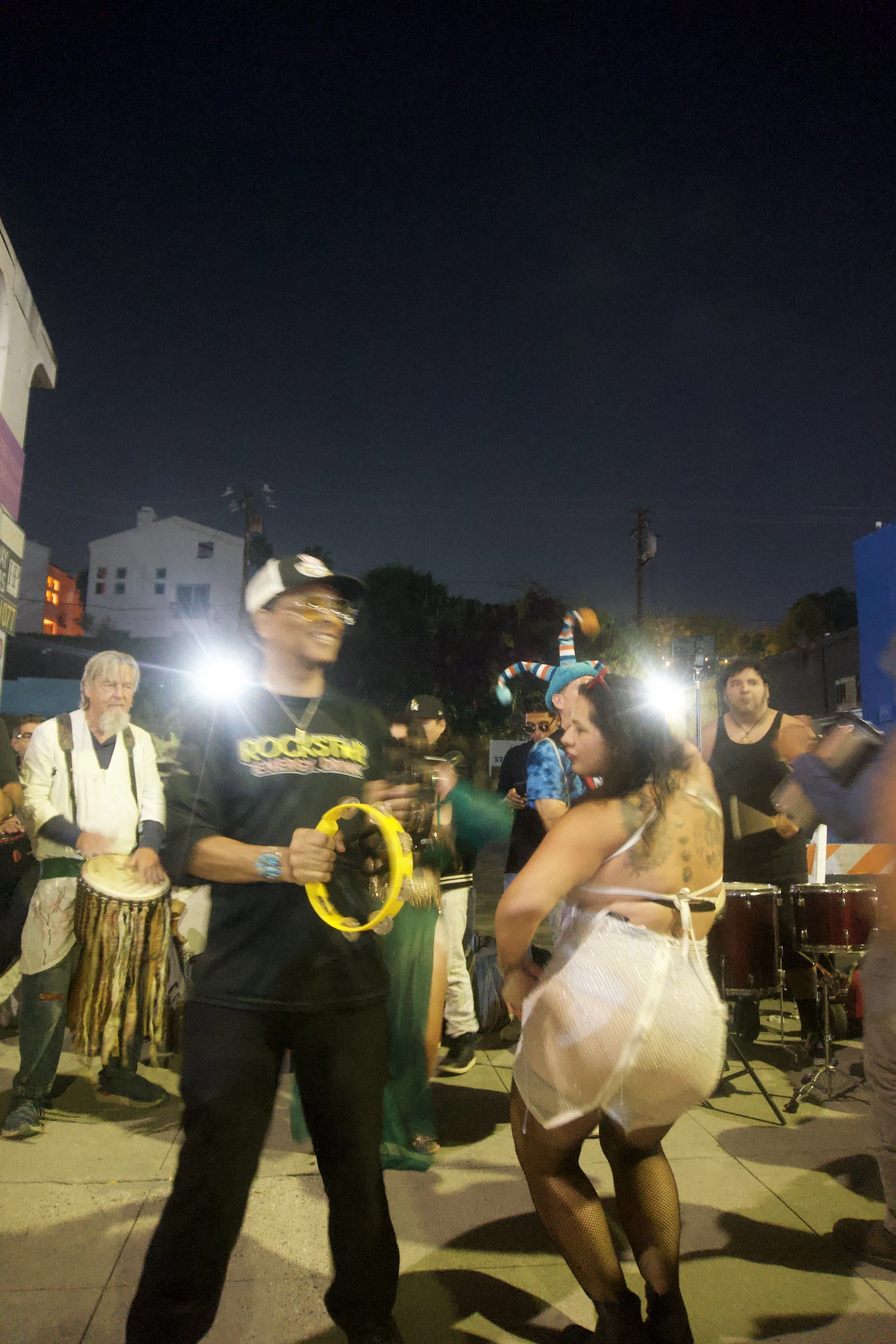 Night street drum circle with people dancing, playing percussion, and celebrating under bright lights, capturing the lively, free-spirited energy of Shy Rockstar Studios.
