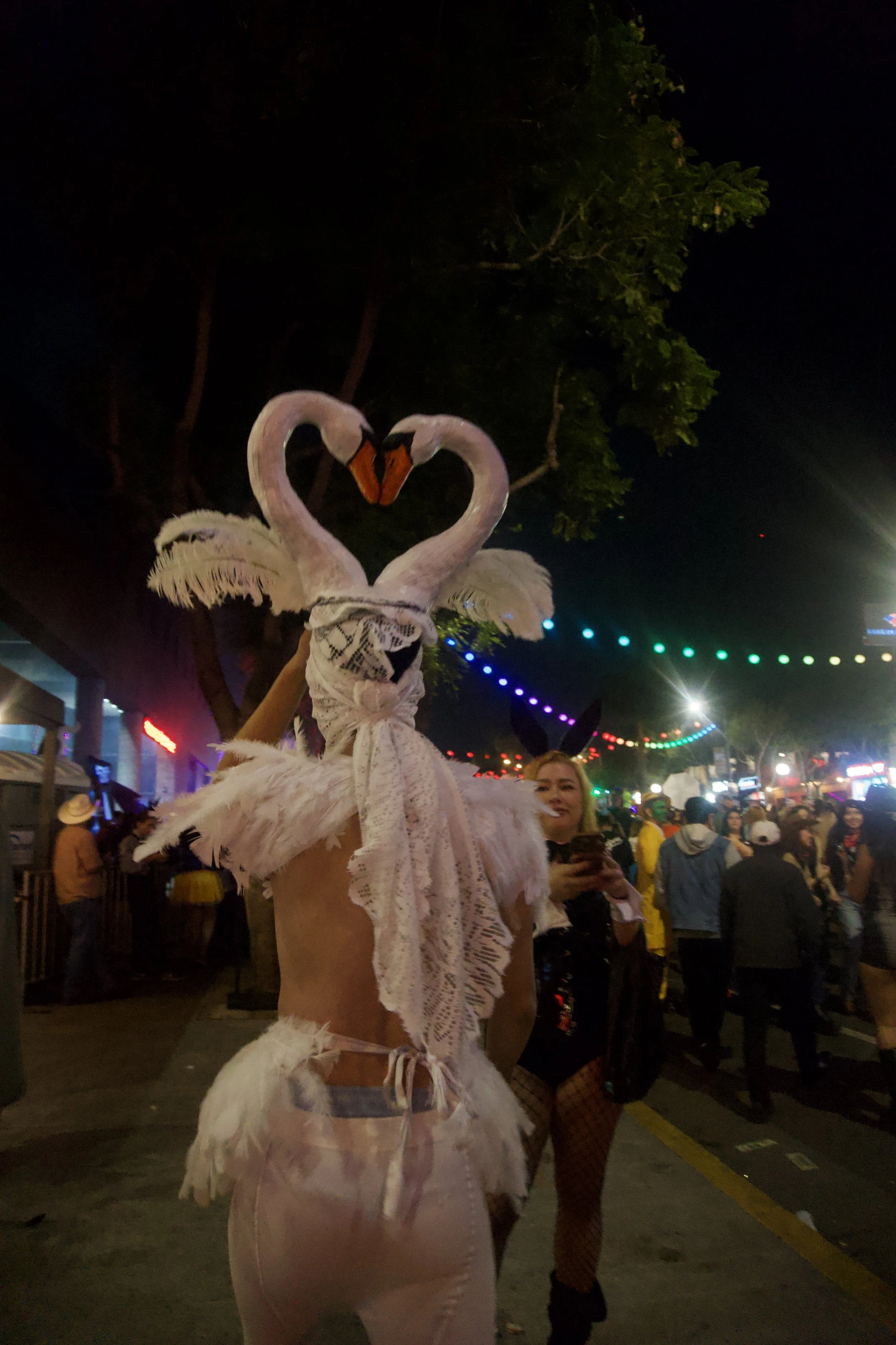 Person wearing an elaborate white swan costume with two swan heads forming a heart shape, standing in a colorful night street festival, capturing the whimsical, surreal energy of Shy Rockstar Studios.