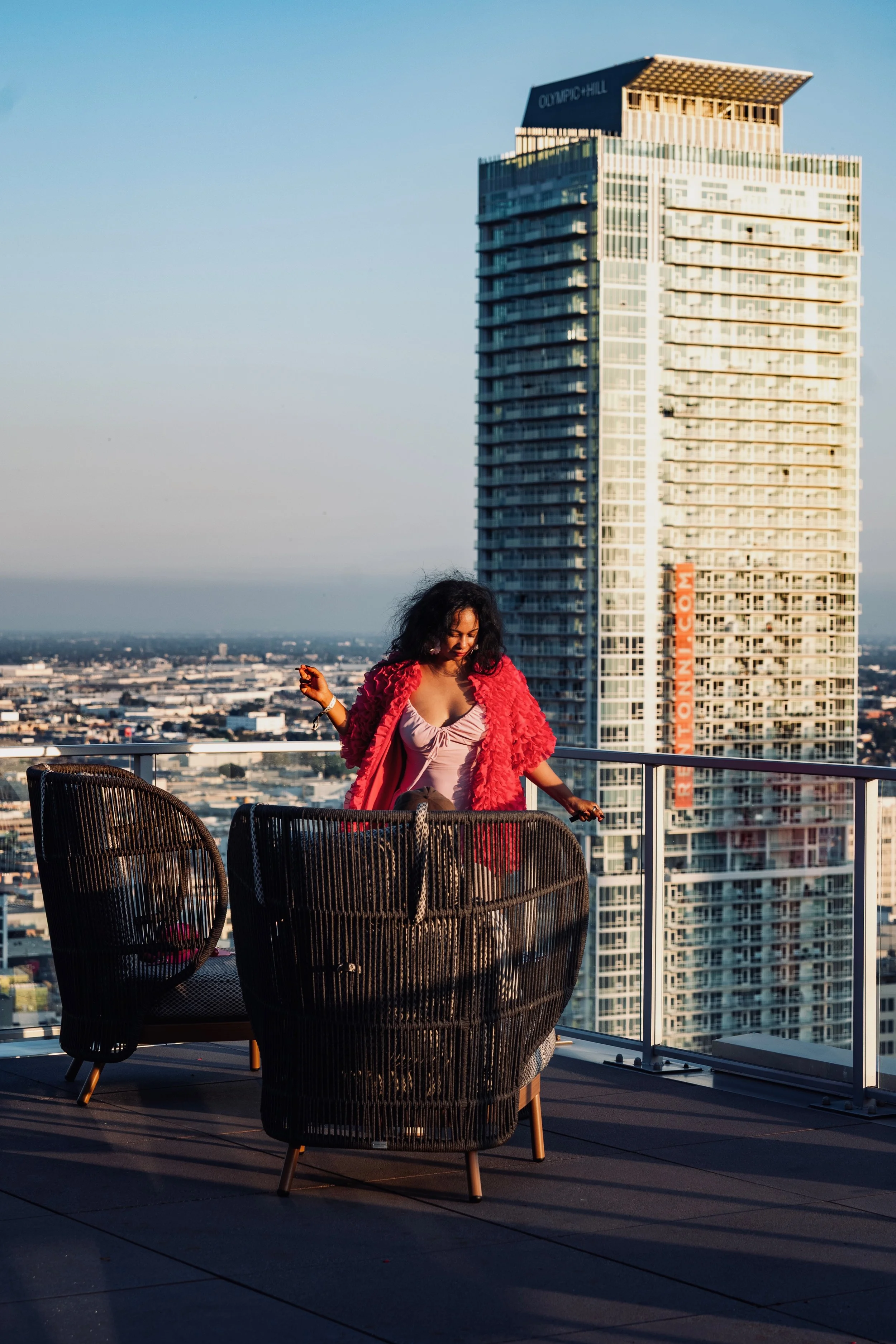 Woman standing on a rooftop at golden hour in a pink top and red textured jacket, overlooking the city skyline, capturing the soft, cinematic glamour of Shy Rockstar Studios.