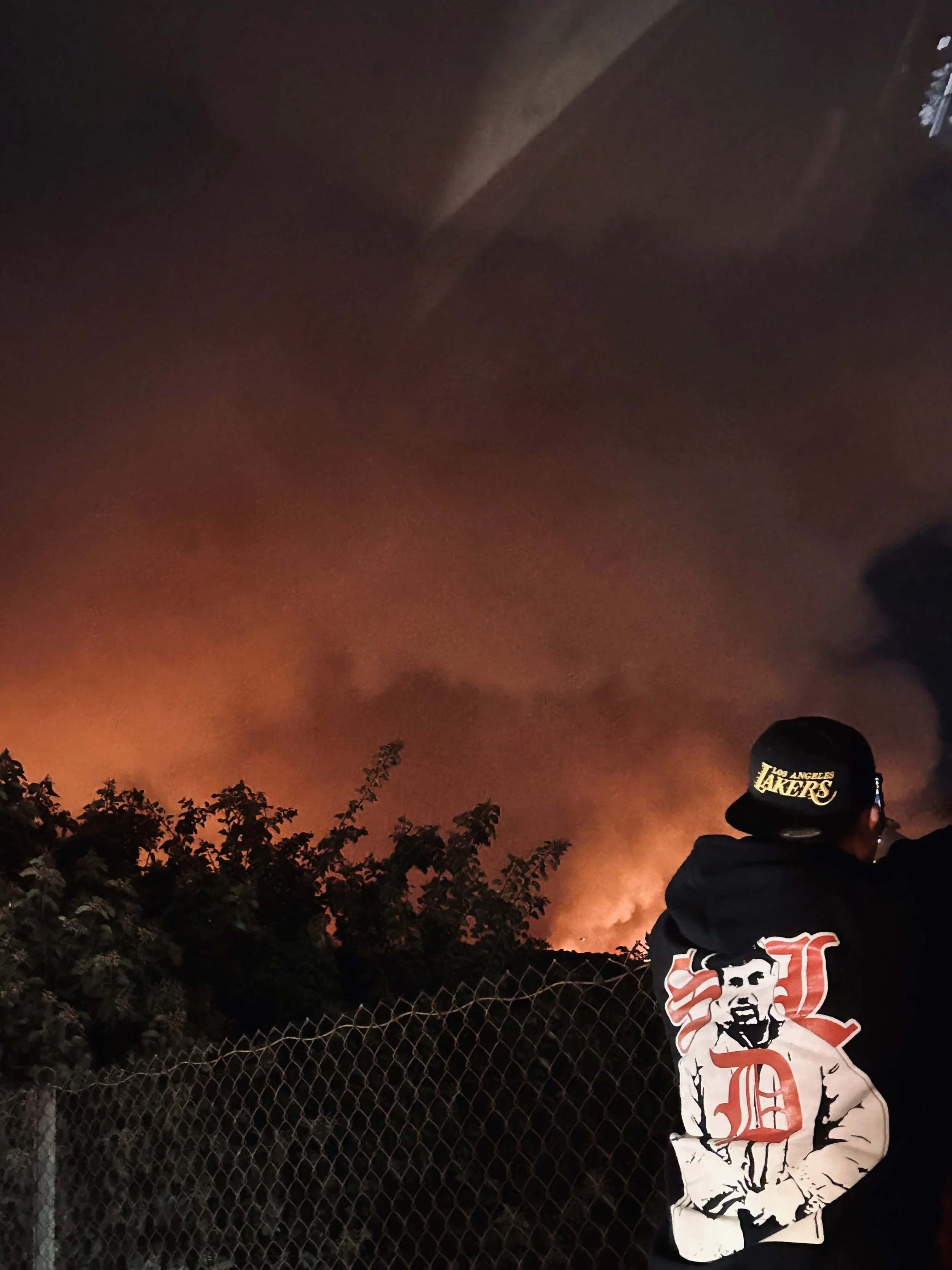Person in Lakers cap watching a wildfire blaze against the night sky.