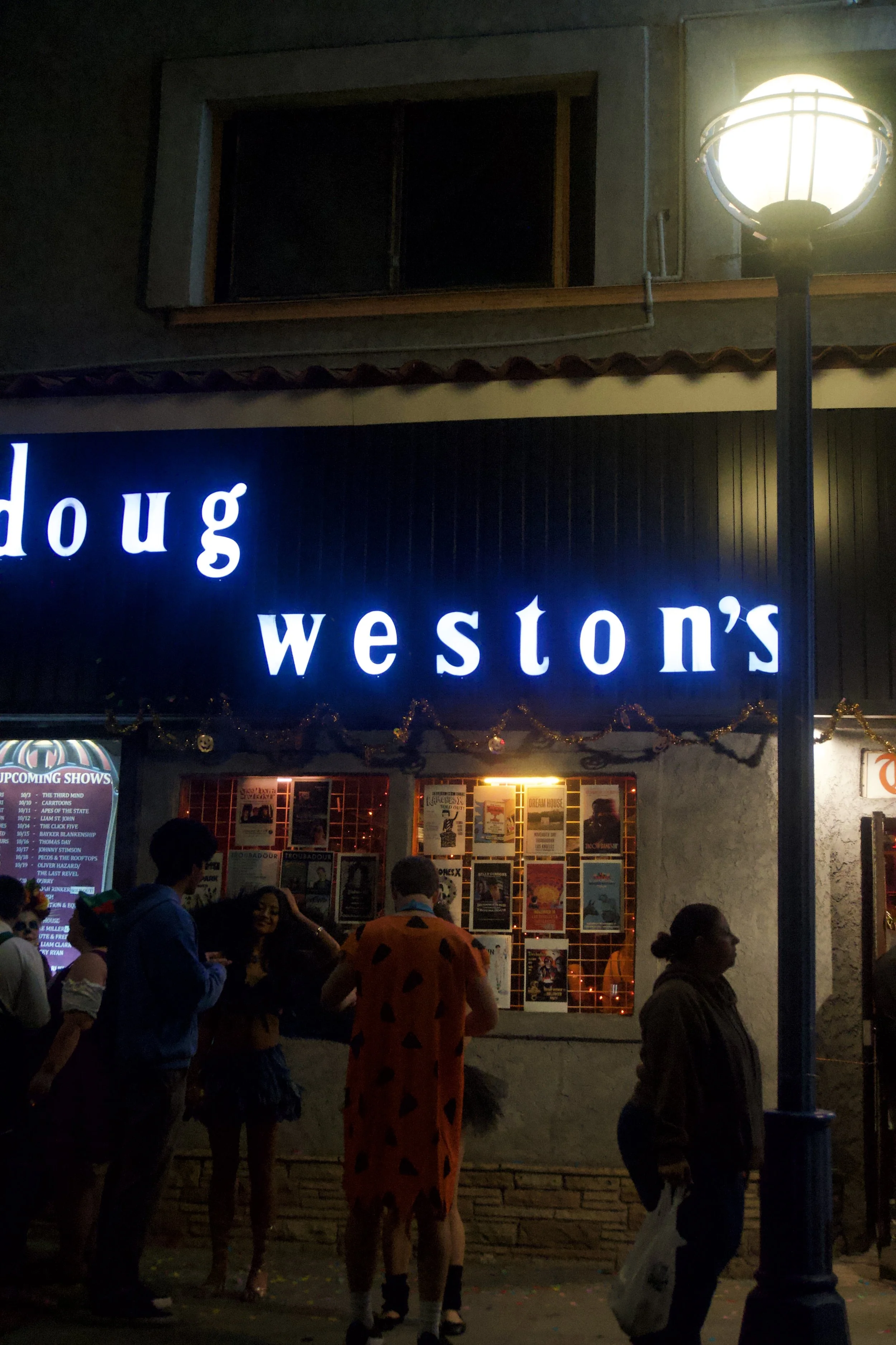 Nighttime street scene outside a venue with a glowing “Doug Weston’s” sign, people in costumes gathering beneath warm window lights, capturing the nostalgic, cinematic energy of Shy Rockstar Studios.