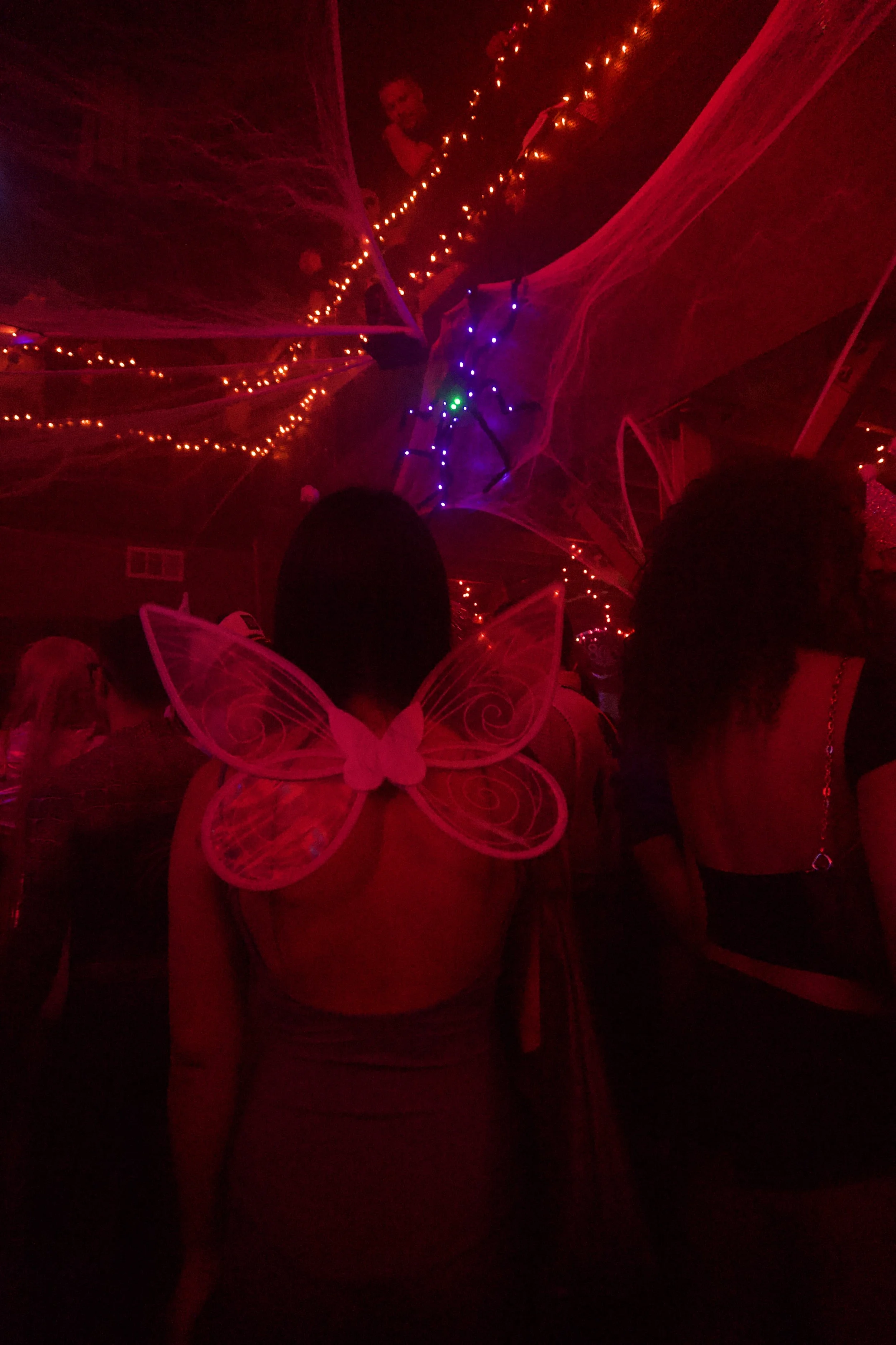 Woman wearing glowing fairy wings in a dim red-lit party filled with webs and string lights, capturing the dreamy, surreal nightlife aesthetic of Shy Rockstar Studios.