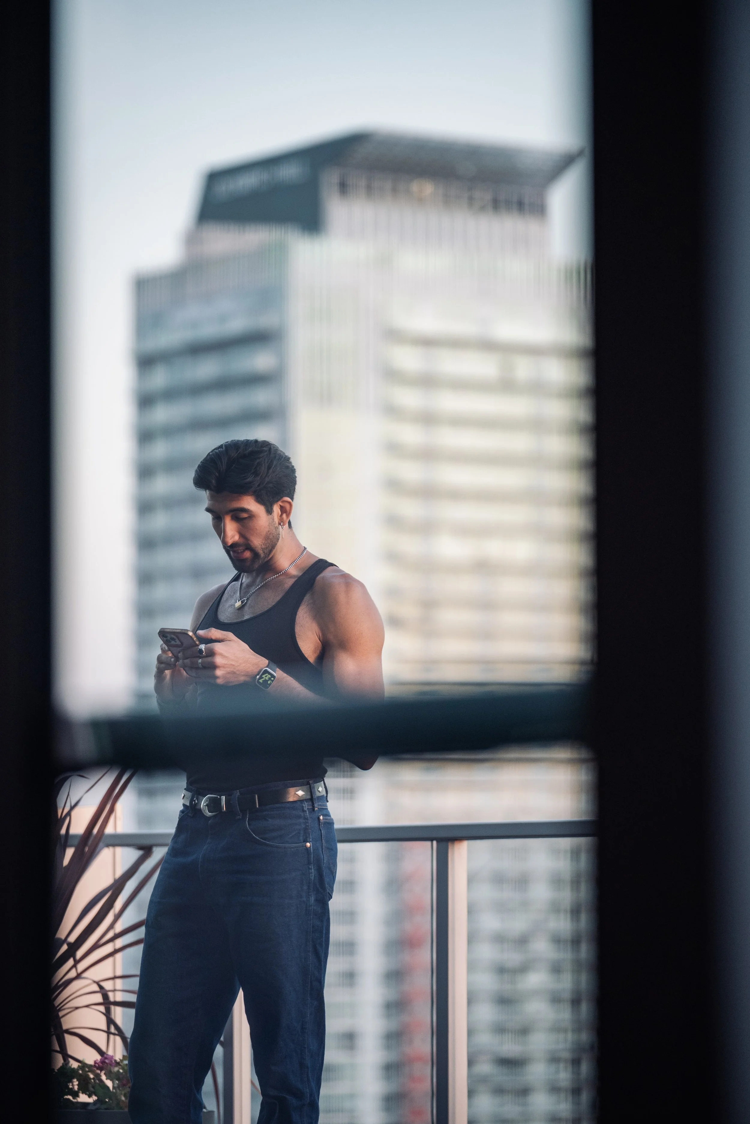 Man standing on a high-rise balcony at golden hour, looking at his phone with the city skyline behind him, capturing the quiet, cinematic mood of Shy Rockstar Studios.