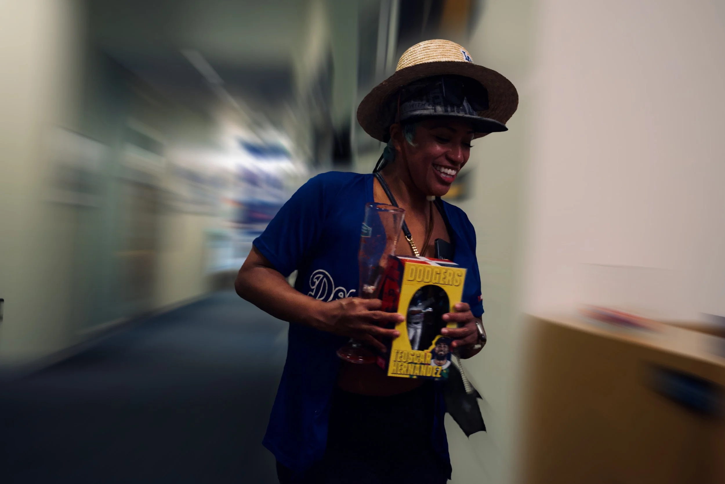 Joyful person in a Dodgers jersey walking through a hallway with motion blur, holding a collectible box and a tall glass, capturing a fun, energetic moment in Shy Rockstar Studios style.