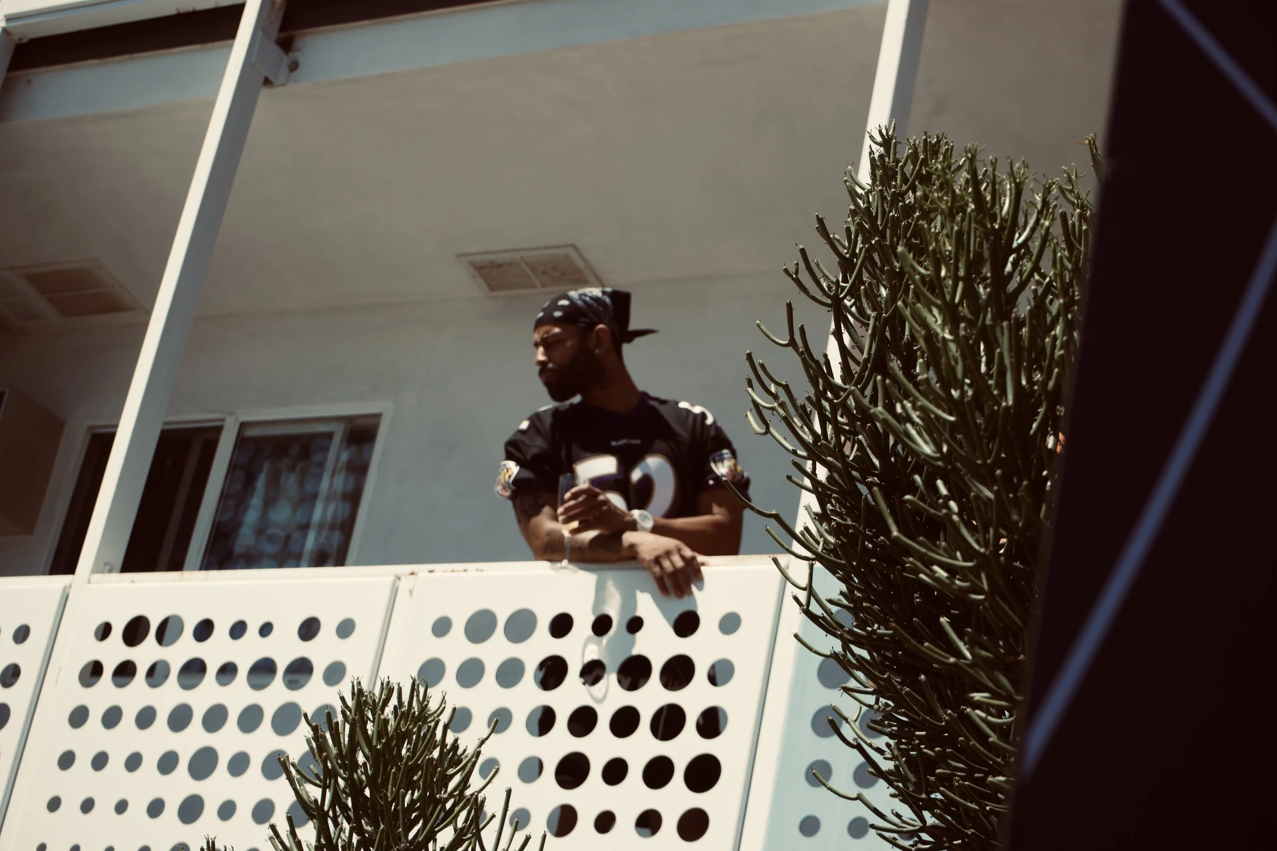 Man in a jersey gazing thoughtfully from a balcony, greenery in the frame.