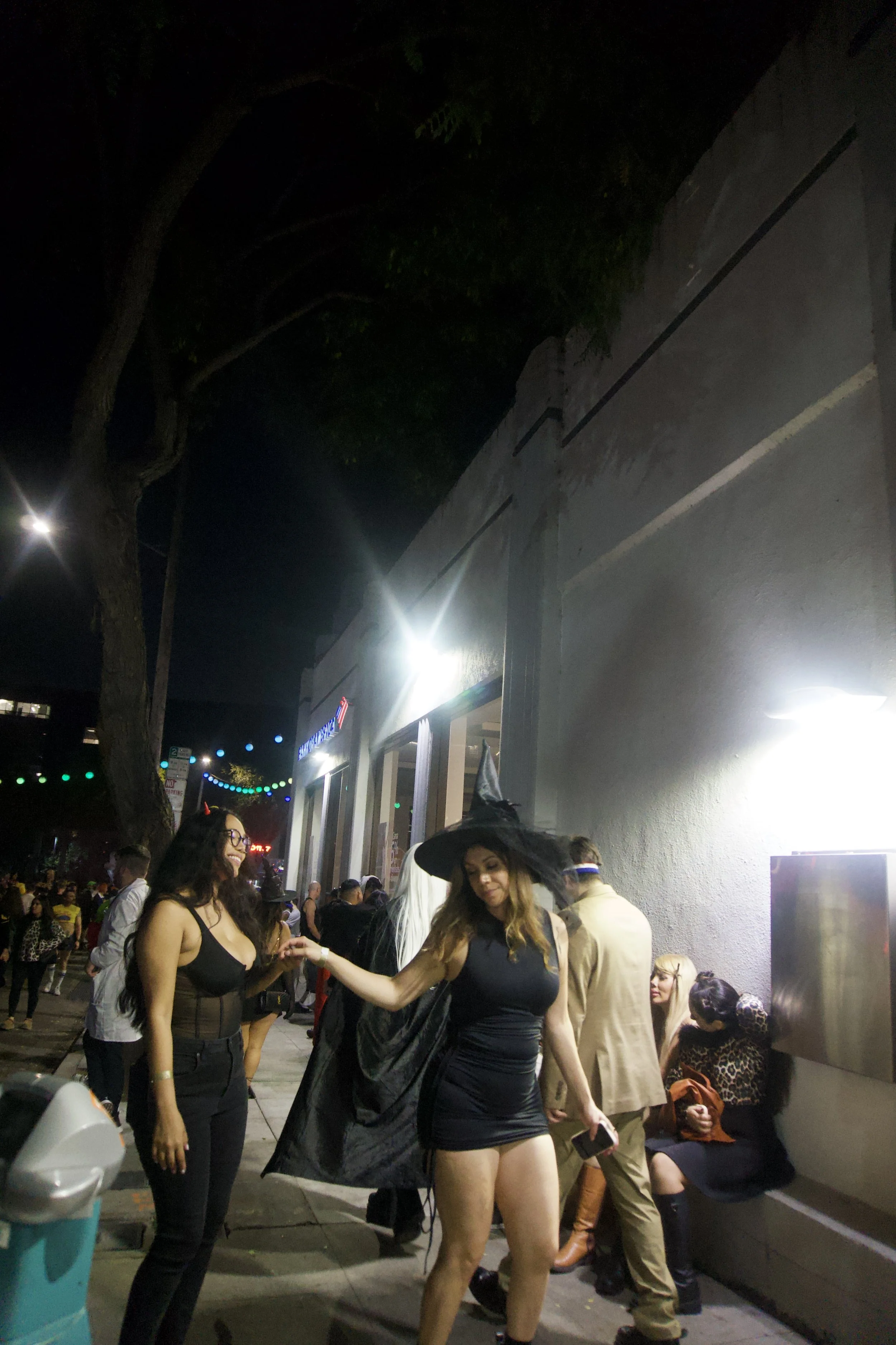 Two women in Halloween costumes chatting on a busy night street lined with bright lights and people, capturing the playful, cinematic energy of Shy Rockstar Studios.