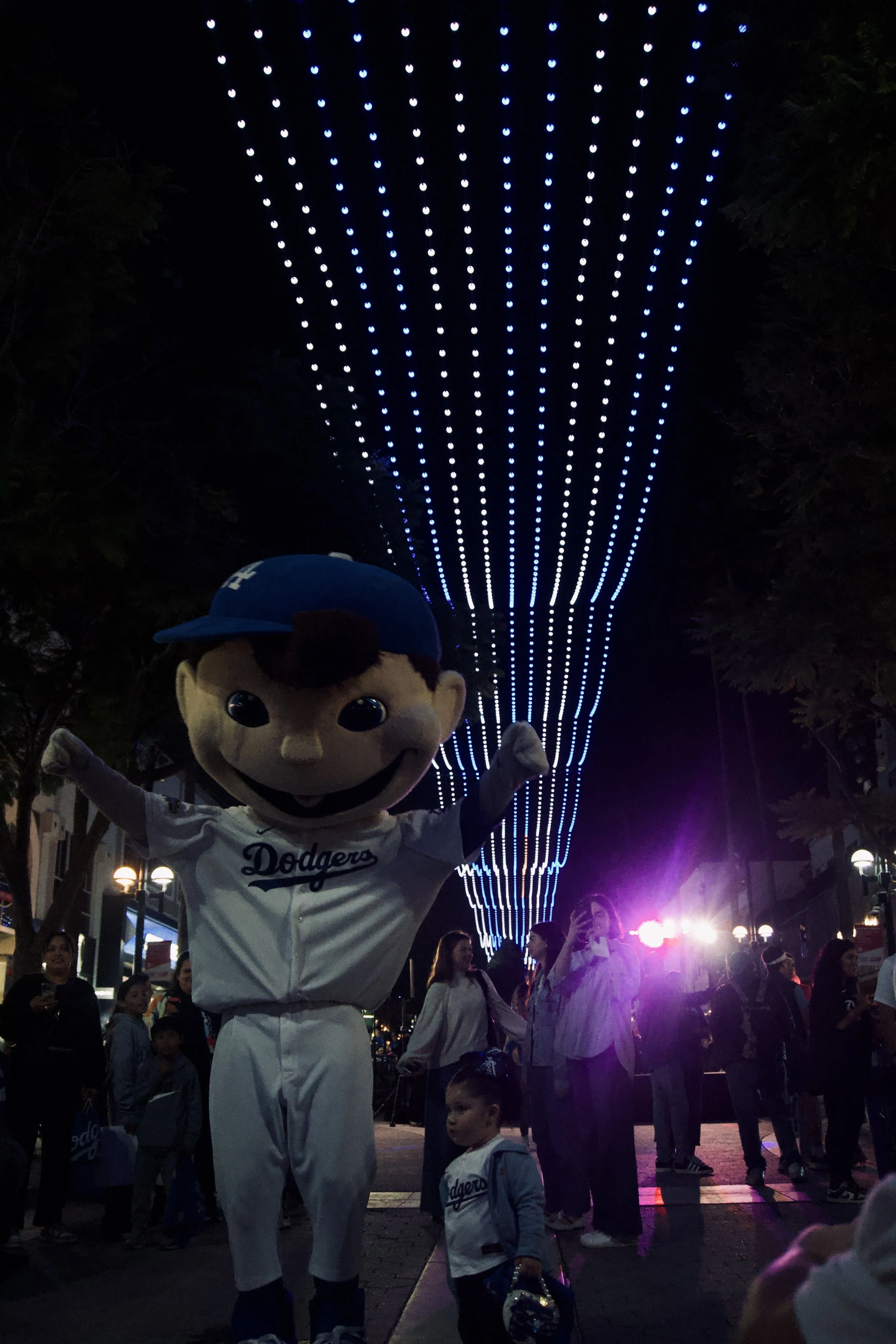 Dodgers mascot posing with arms raised under a tunnel of glowing blue lights during a lively night celebration, surrounded by a crowd, capturing the playful cinematic energy of Shy Rockstar Studios.