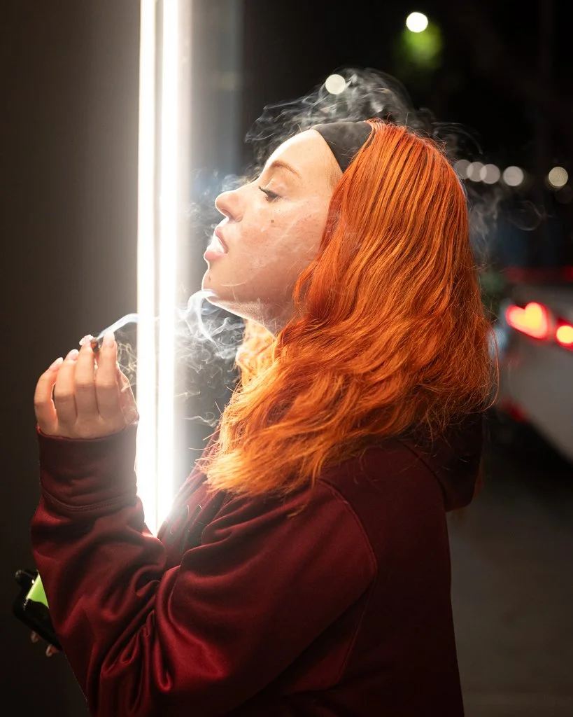 Woman with vibrant orange hair exhaling smoke beside a bright vertical light, creating a dreamy, cinematic glow in true Shy Rockstar Studios style.