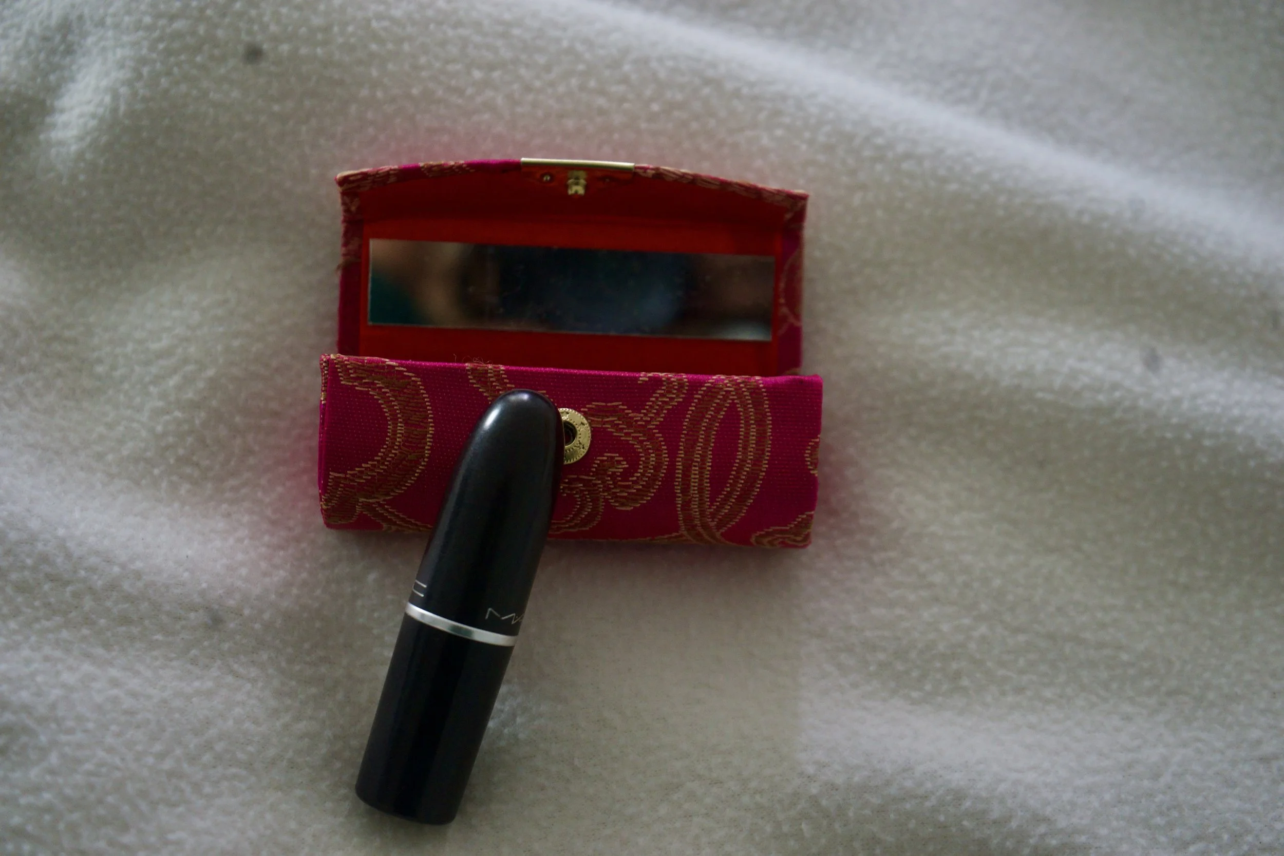 A black lipstick stands upright before a small red patterned case with a partially revealed mirror, centered against soft white fabric, composed with restrained framing and intimate depth to suggest ritual and calibration