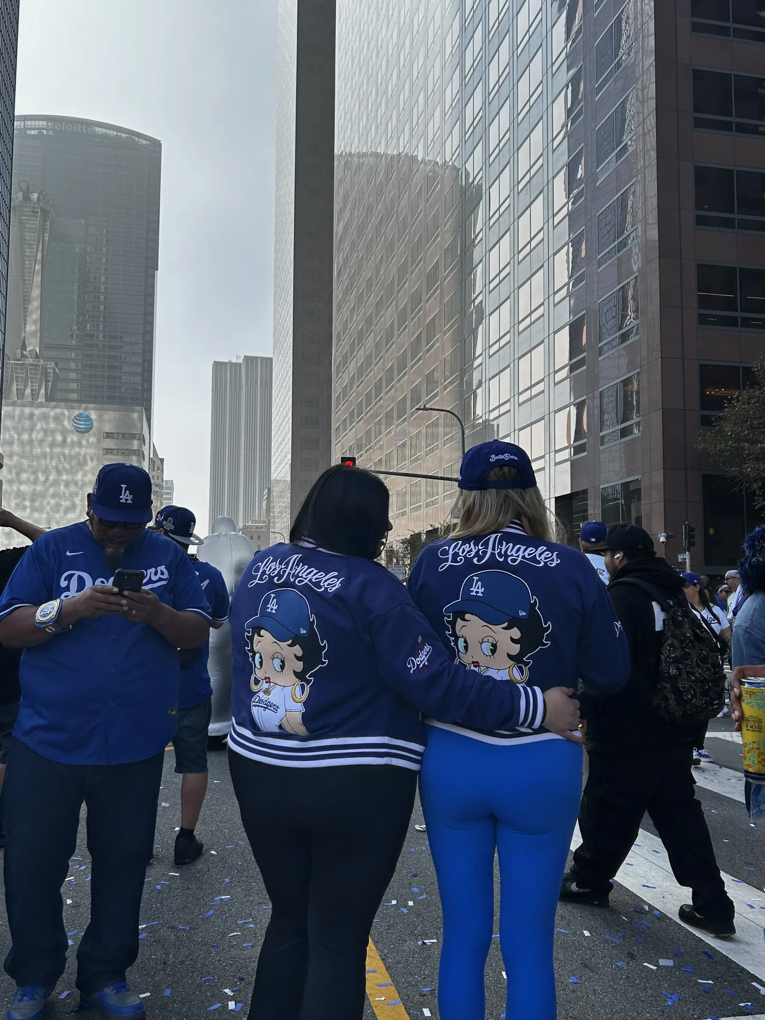 Two people wearing matching Los Angeles Dodgers jackets with cartoon designs, walking together through a crowded downtown street, capturing a warm, nostalgic city moment in Shy Rockstar Studios style.