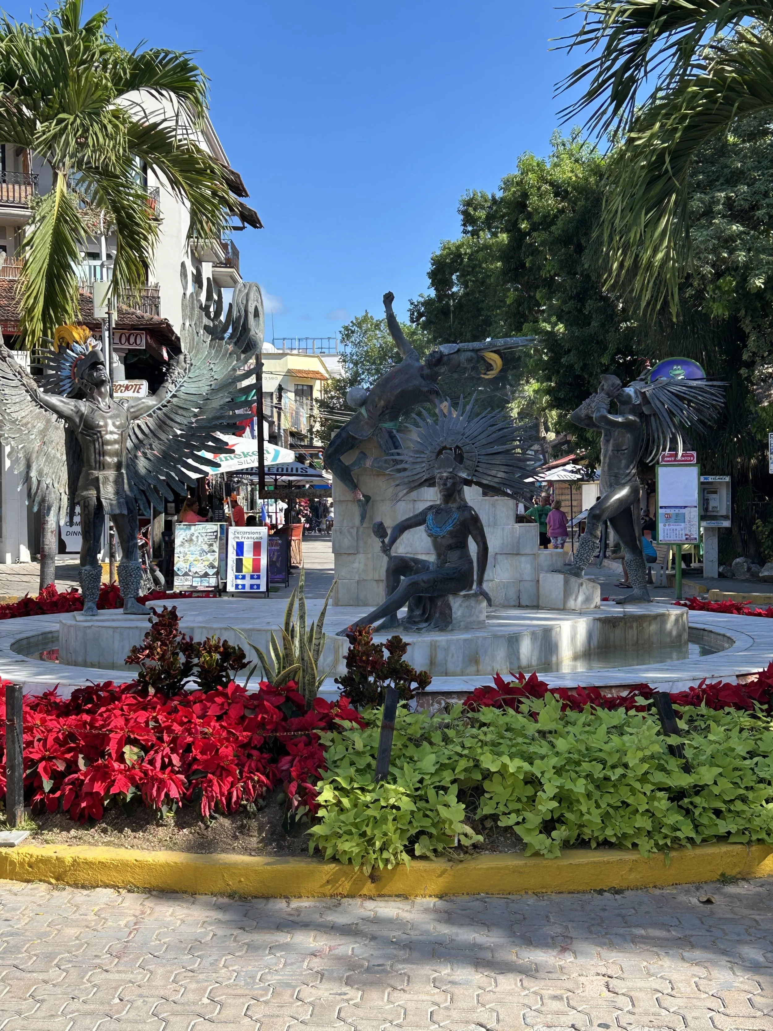 Bronze sculptures depicting indigenous figures with feathered headdresses in an outdoor plaza, surrounded by red and green foliage under palm trees.