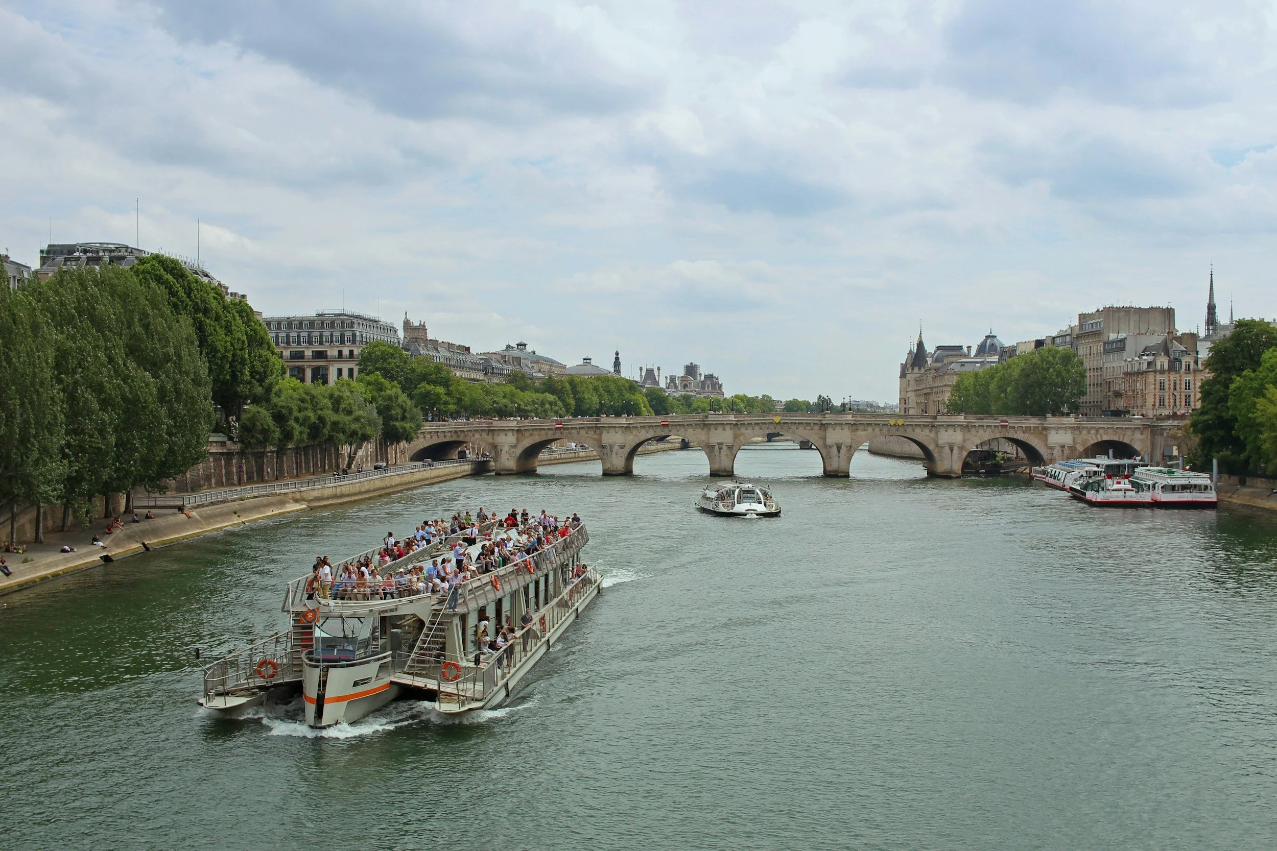 A scenic view of the Seine River in Paris with a boat filled with tourists, other boats on the river, and historic buildings and bridges along the riverbank under a partly cloudy sky.