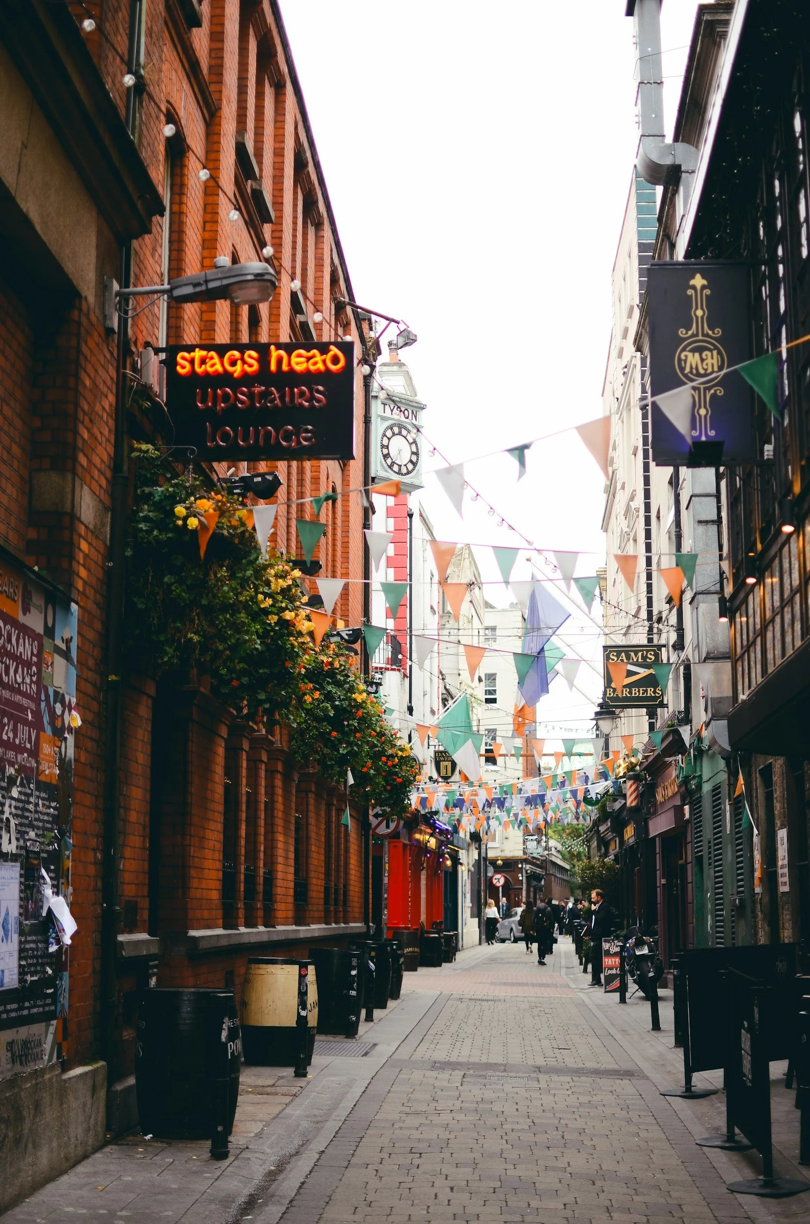 A narrow city street decorated with colorful triangular flags hanging across the street, lined with brick and stone buildings featuring signs for bars and lounges, with pedestrians walking along the sidewalk.