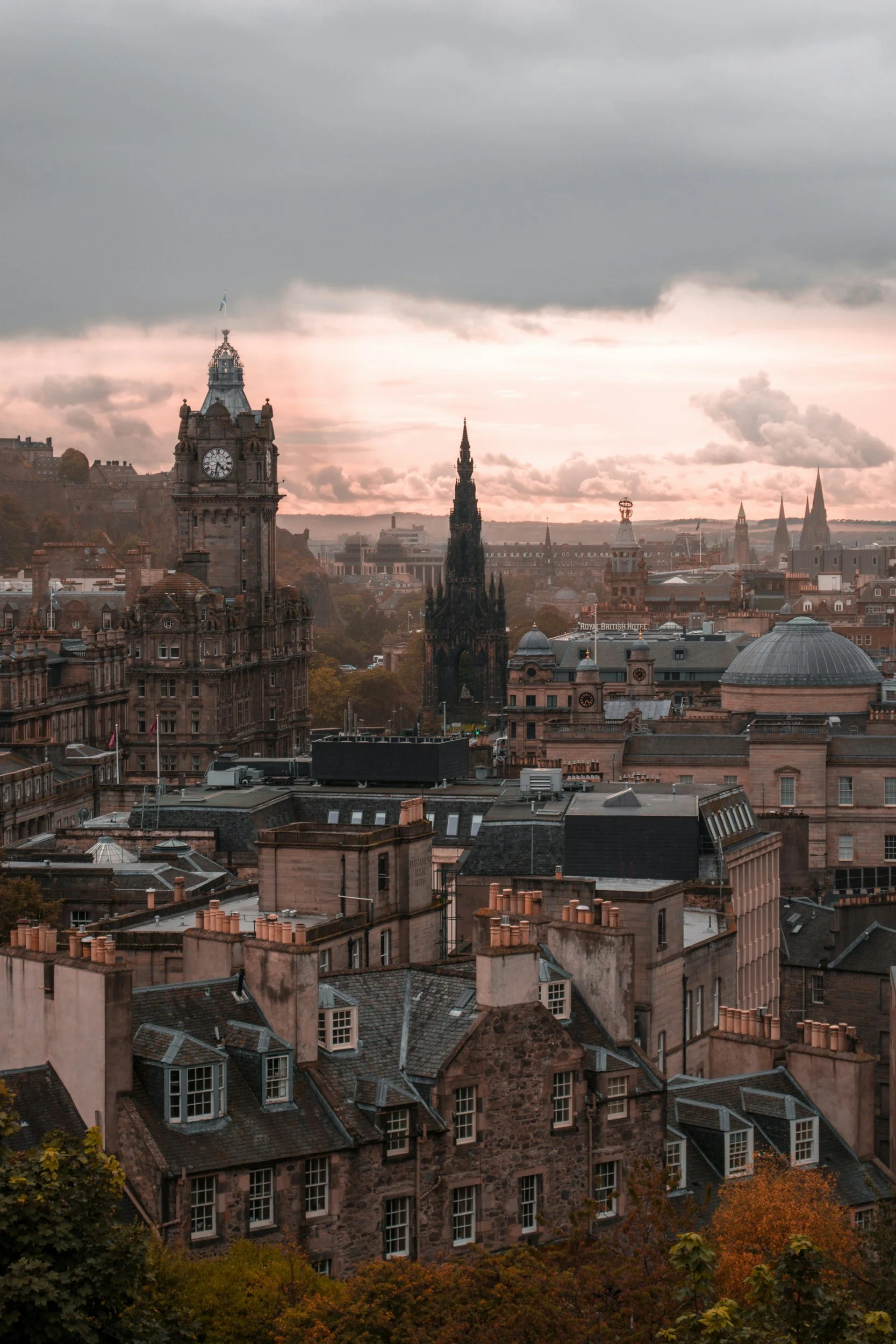 A city skyline during sunset with historic buildings and spires, including a clock tower.