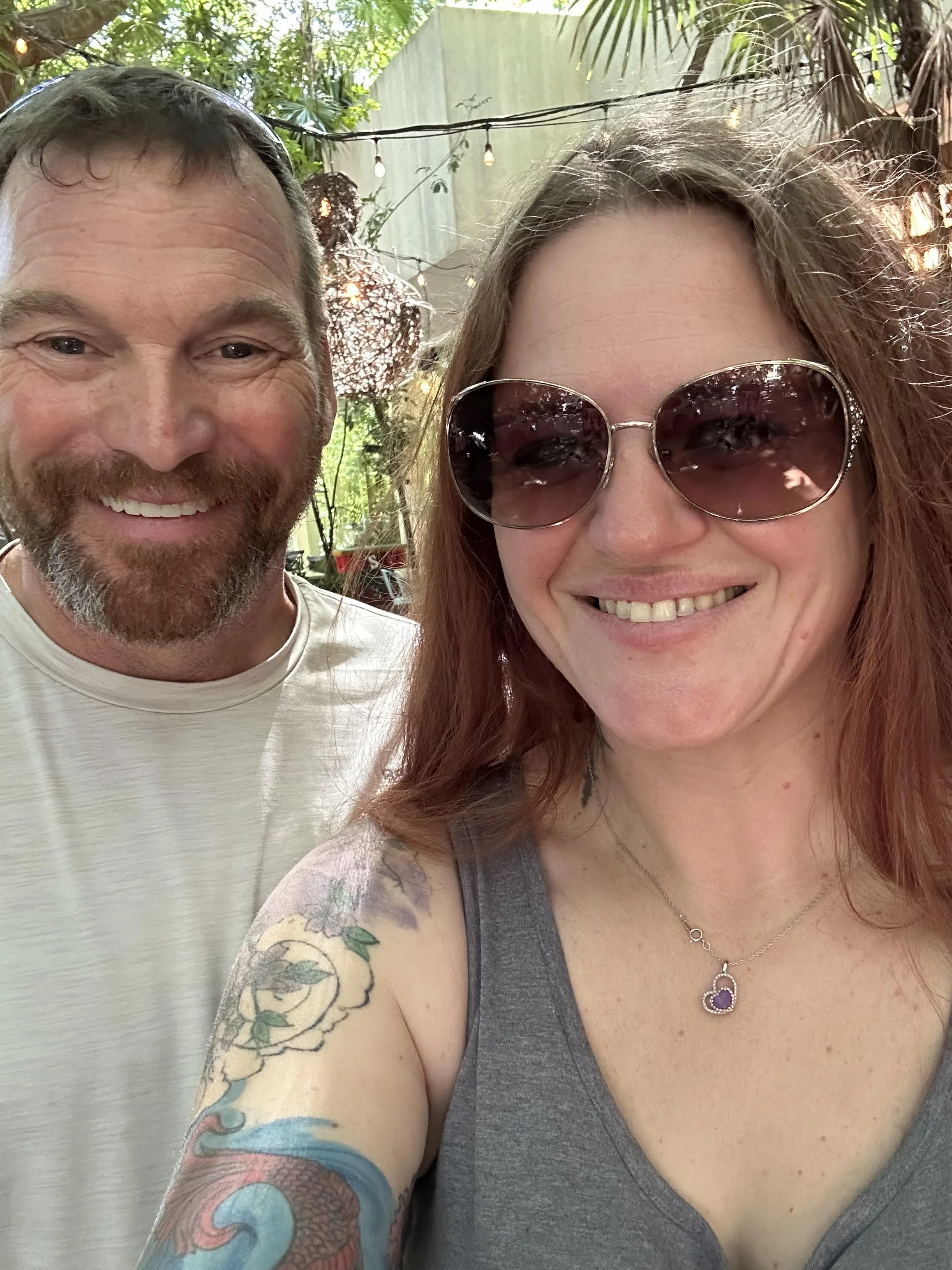 A smiling man with a beard and a woman with red hair, sunglasses, and a tattoo on her arm taking a selfie outdoors in a garden setting with string lights and greenery.