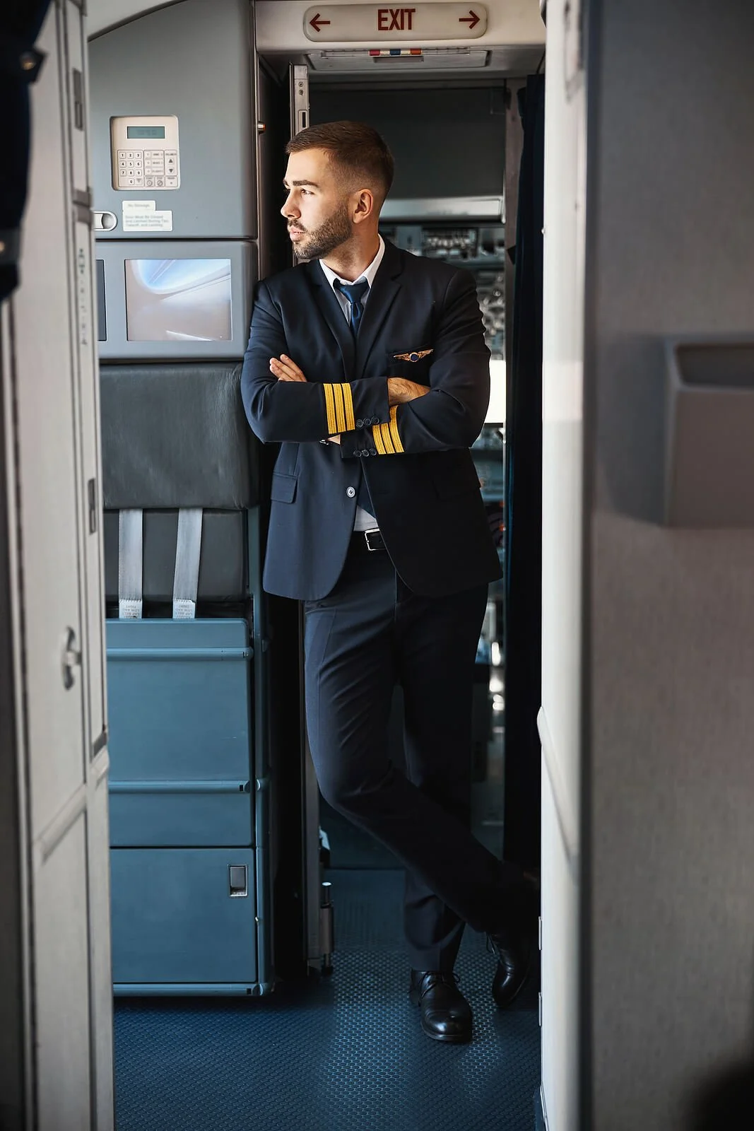 Commercial airline pilot in uniform with arms crossed standing in aircraft cabin near exit sign. Understand how irregular schedules impact your health through expert SAP evaluations in Las Vegas, NV.