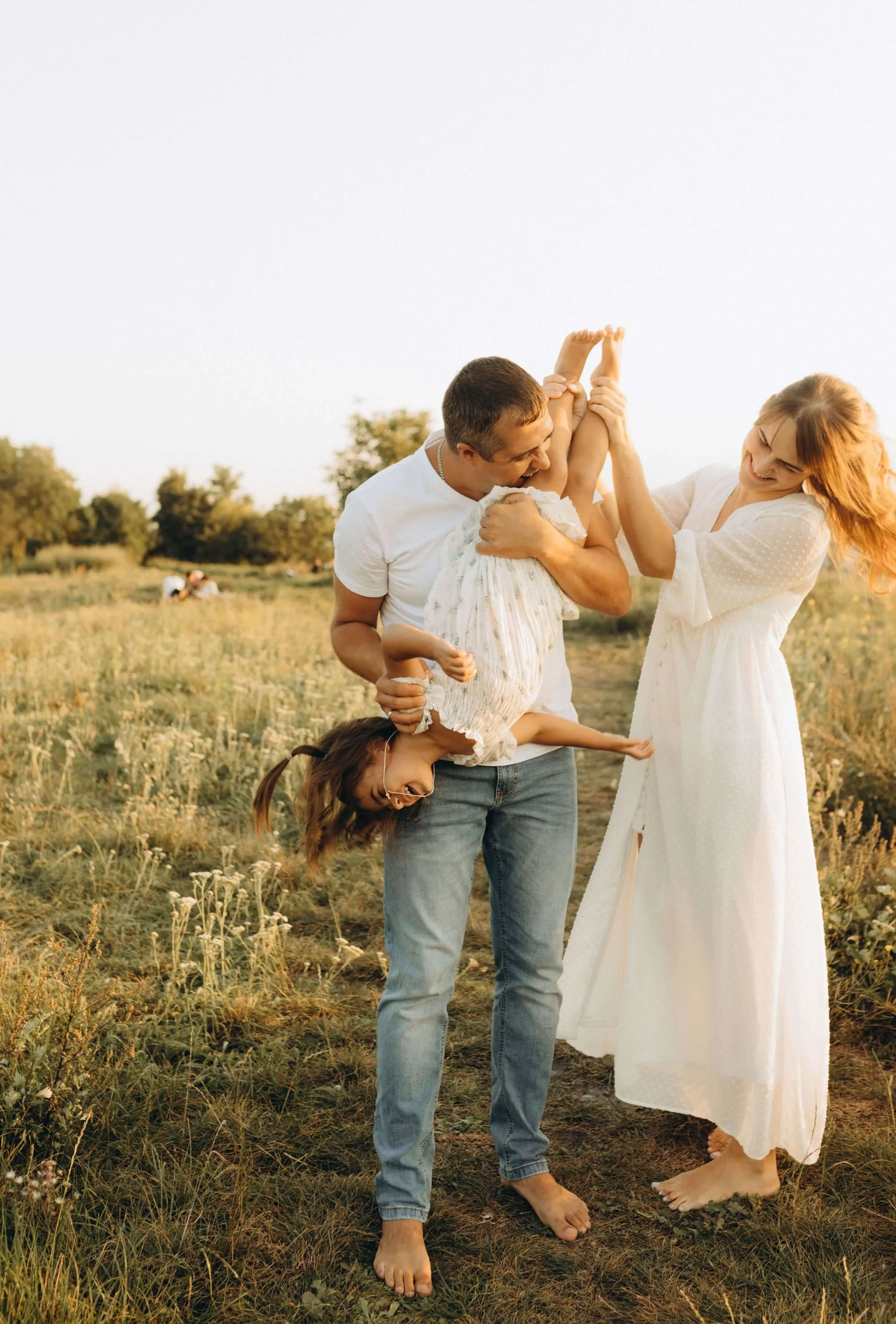 Two parents laughing and playfully flipping a young child upside down in an open grassy field at sunset. Parenting therapy in Los Angeles, CA helps you build the skills and confidence to show up more fully in the moments that matter most.