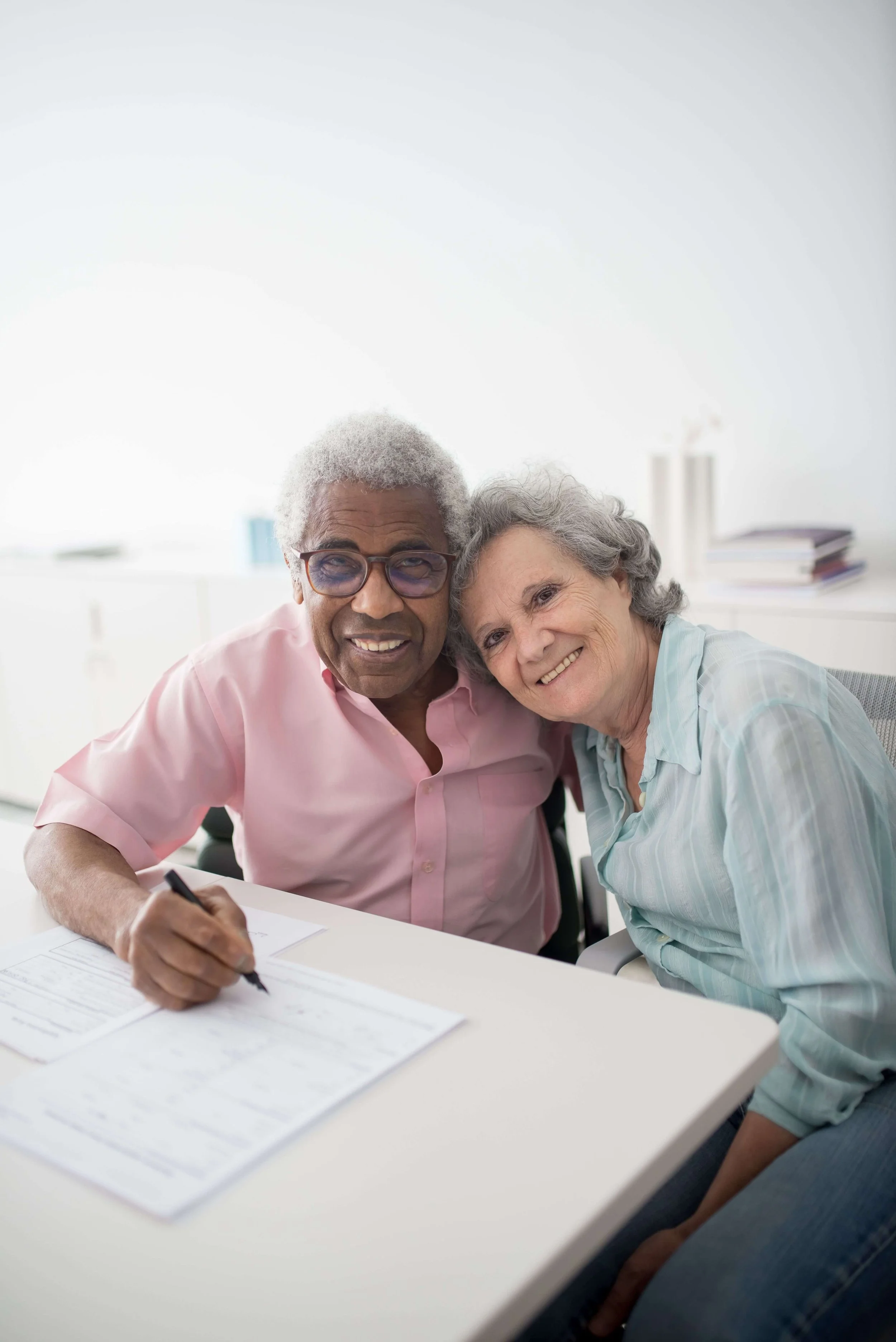 An older couple smiling warmly at the camera while seated at a desk with paperwork in a bright room. Life care plans in Los Angeles, CA are designed to give individuals and their families a clear, confident path forward after injury or trauma.