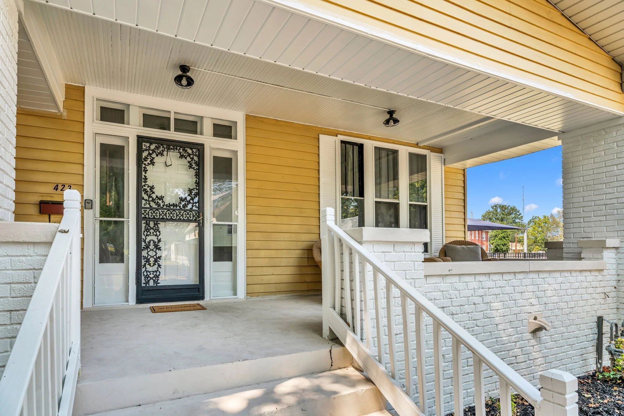 Front porch of a yellow house with white brick accents, black decorative security door, white railing, and windows with white shutters, under a covered roof with black ceiling lights, and a sunny blue sky background.