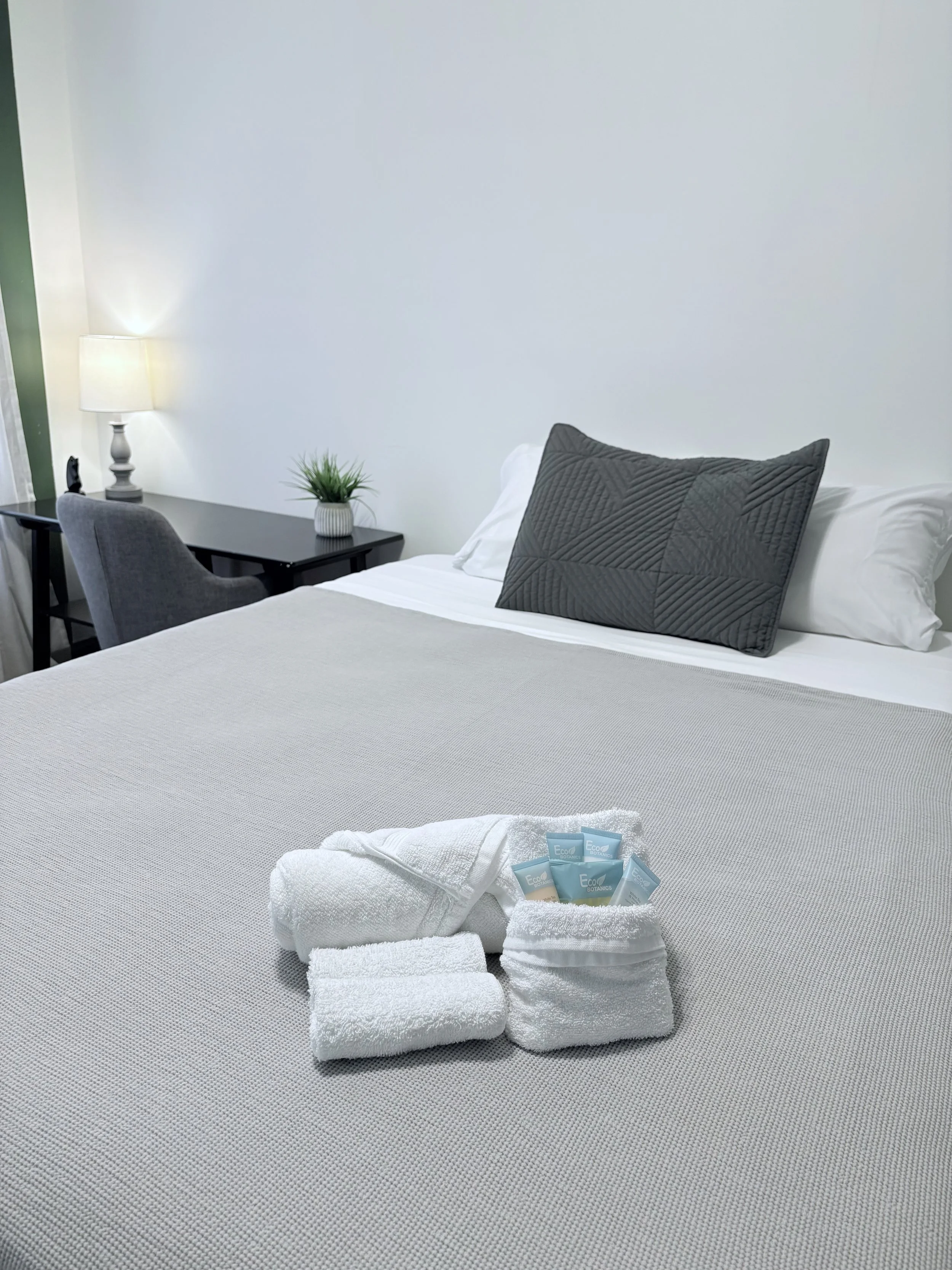 Clean hotel bed with rolled towels, toiletries, and a decorative pillow, in a modern, minimalistic room with a side table, lamp, and a plant.