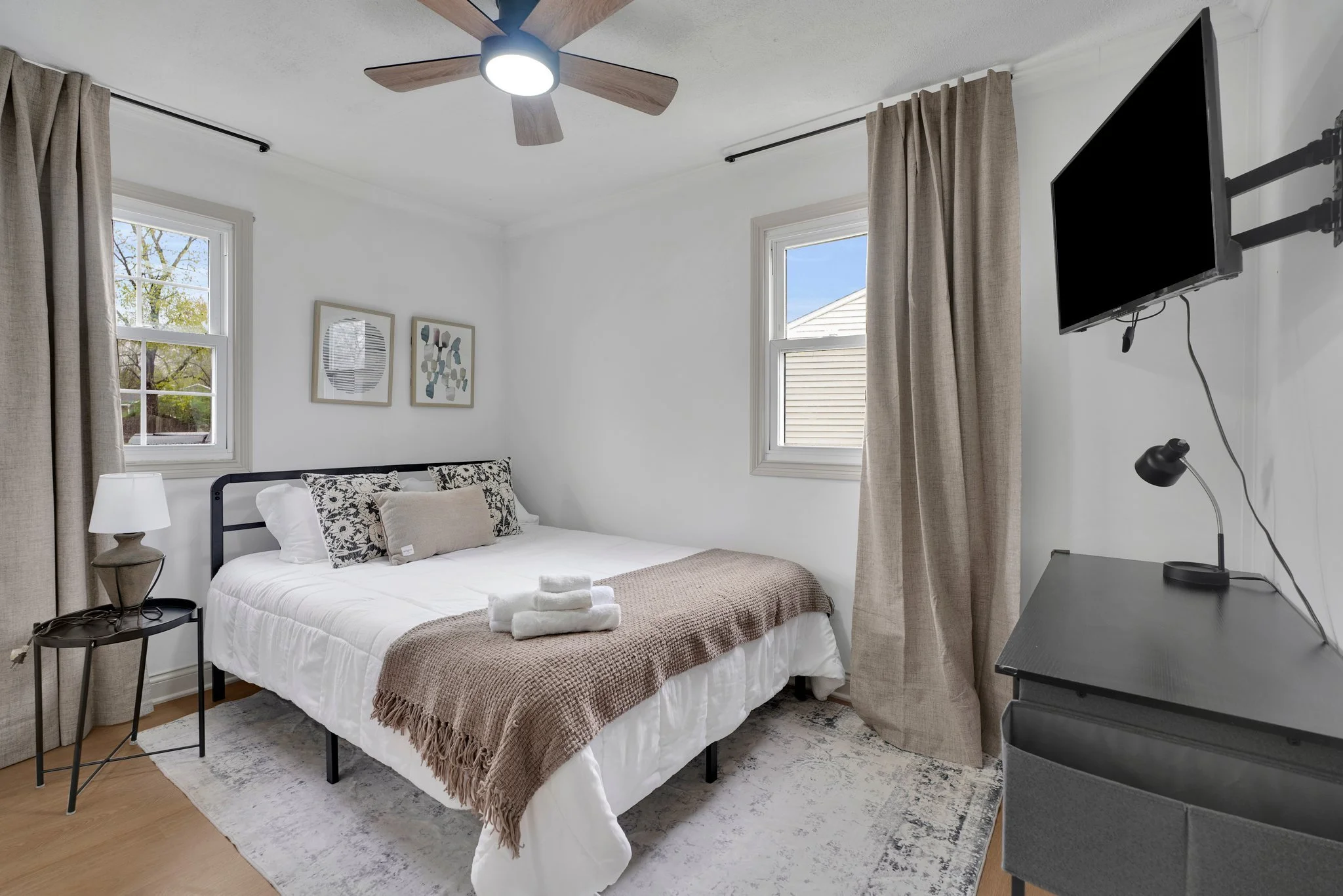 A bedroom with a white bed, beige blanket, and decorative pillows, two windows with beige curtains, a ceiling fan, a side table with a lamp, a wall-mounted TV, and artwork above the bed.