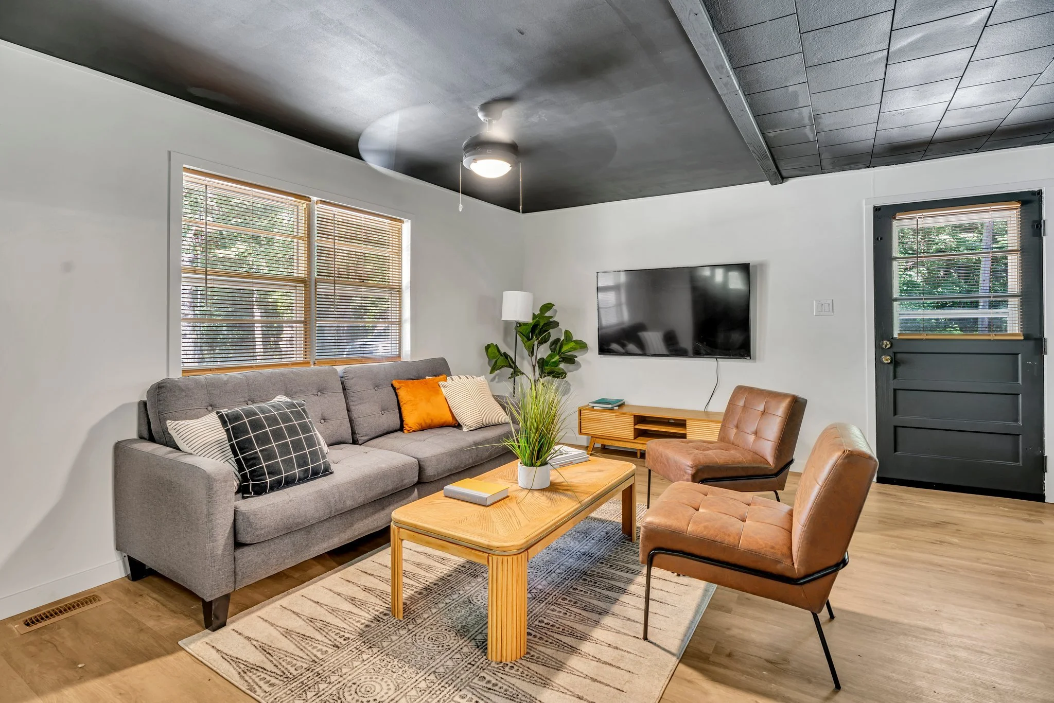 Living room with a gray sofa, two brown chairs, a wooden coffee table, and a flat-screen TV on the wall. There are large windows with blinds, and a door with a window to the outside.