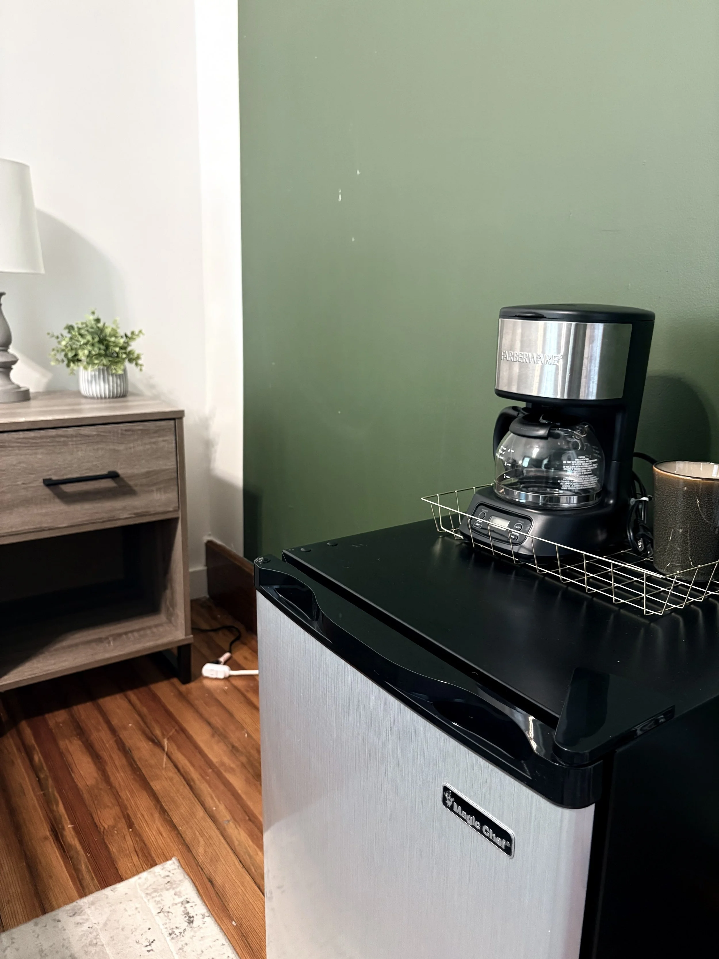 Coffee maker sitting on top of a mini fridge in a room with wooden floor and green wall. A small wooden table with a white lamp and potted plant is in the background.