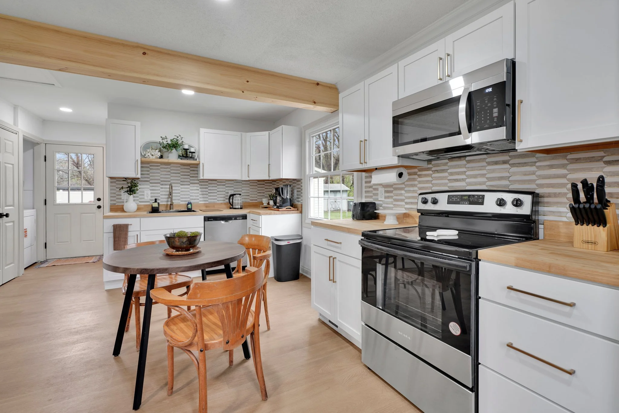 Modern white kitchen with wooden accents, stainless steel appliances, and a small round dining table with wooden chairs.