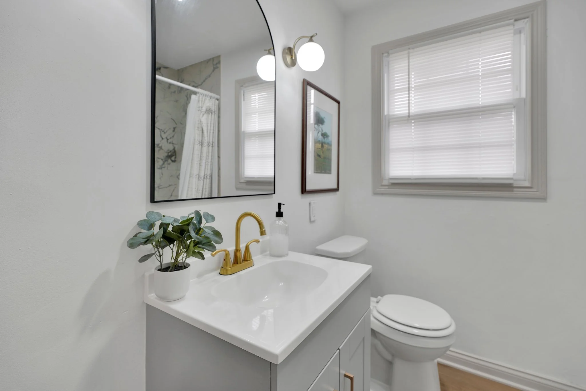 Bathroom with white walls, a mirror, a window with blinds, a small plant on the sink, a gold faucet, a soap dispenser, a framed picture on the wall, a toilet, and a light fixture with two round bulbs.