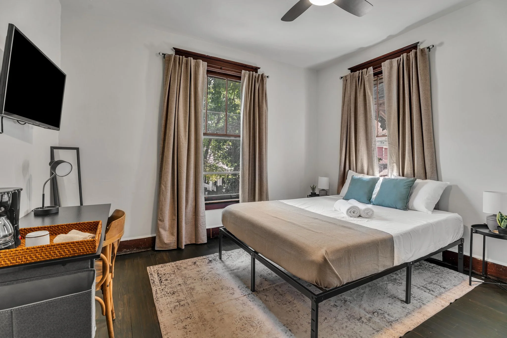 A bedroom with a bed, two windows with beige curtains, a TV on the wall, and a small desk with a lamp and coffee supplies.