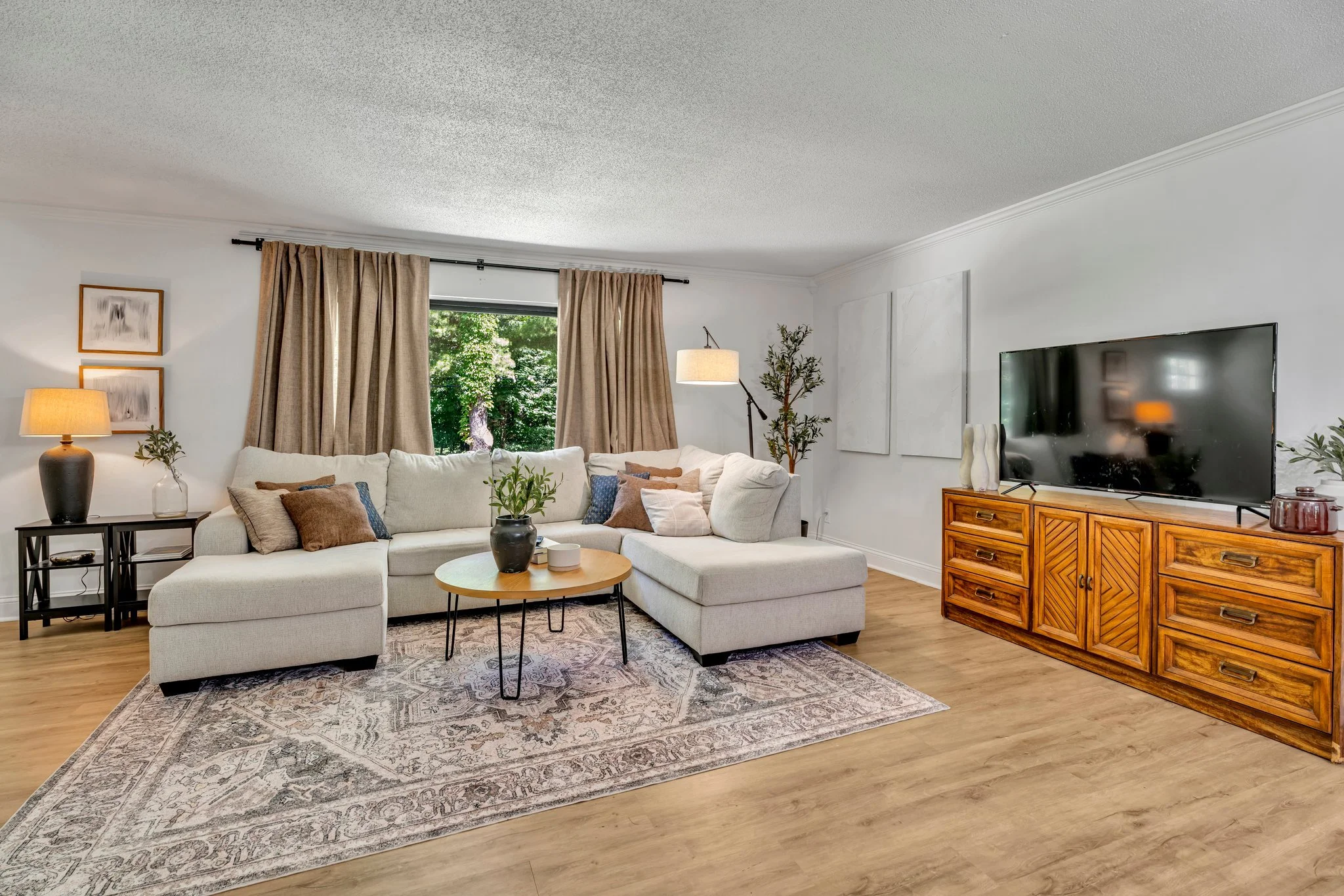 A living room with a large white sofa, beige curtains, a TV on a wooden stand, a potted plant, and various decorative pillows and lamps.