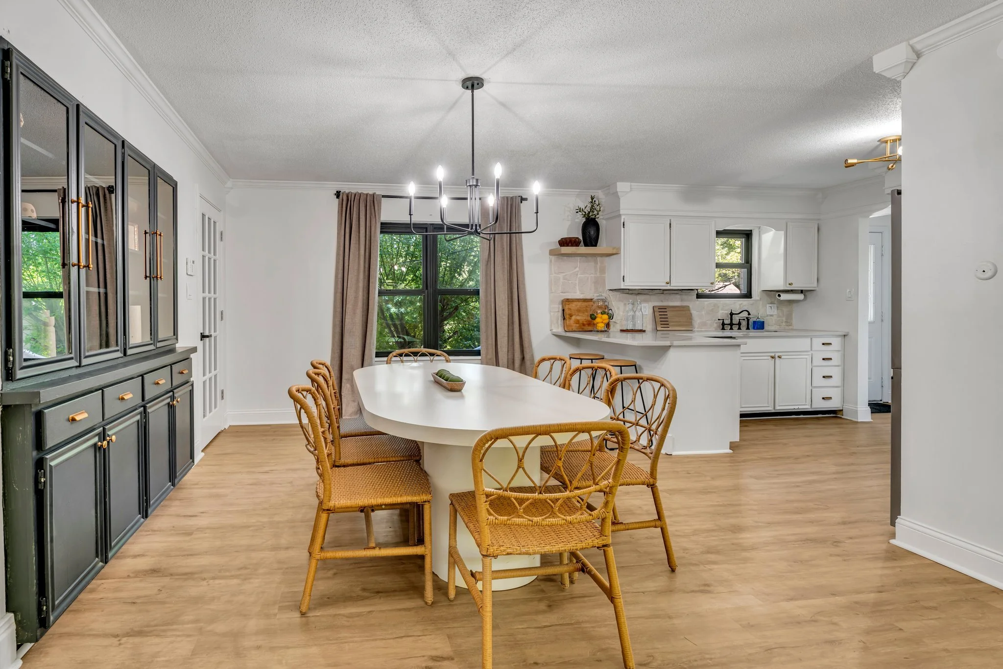 Dining room with white oval table, six rattan chairs, black chandelier, large window with brown curtains, white kitchen in background with black faucet, wooden shelves, and decor.