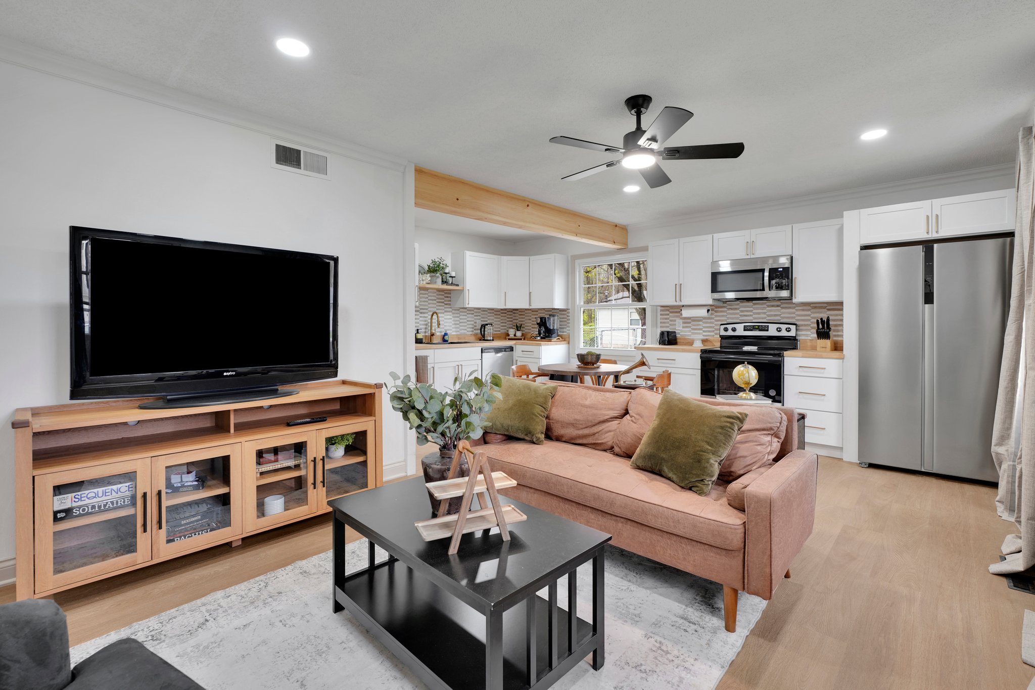 Open-concept living room and kitchen with white cabinets, stainless steel appliances, a beige sofa, a black coffee table with decor, and a large flat-screen TV on a wooden stand.