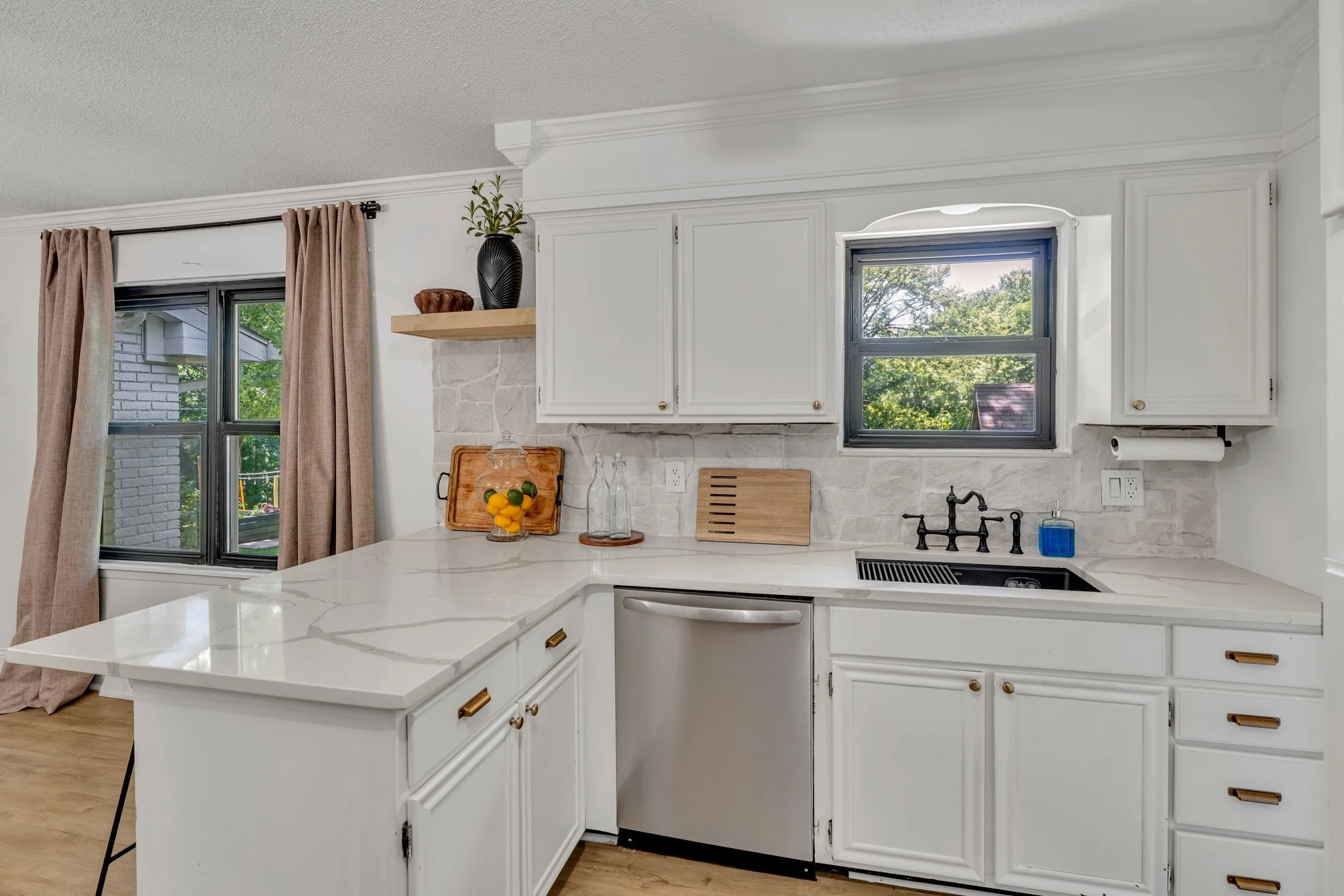 Kitchen with white cabinets, a window above the sink, black faucet, and light-colored countertops. There are decorative items on the counter, and beige curtains on a window to the left.