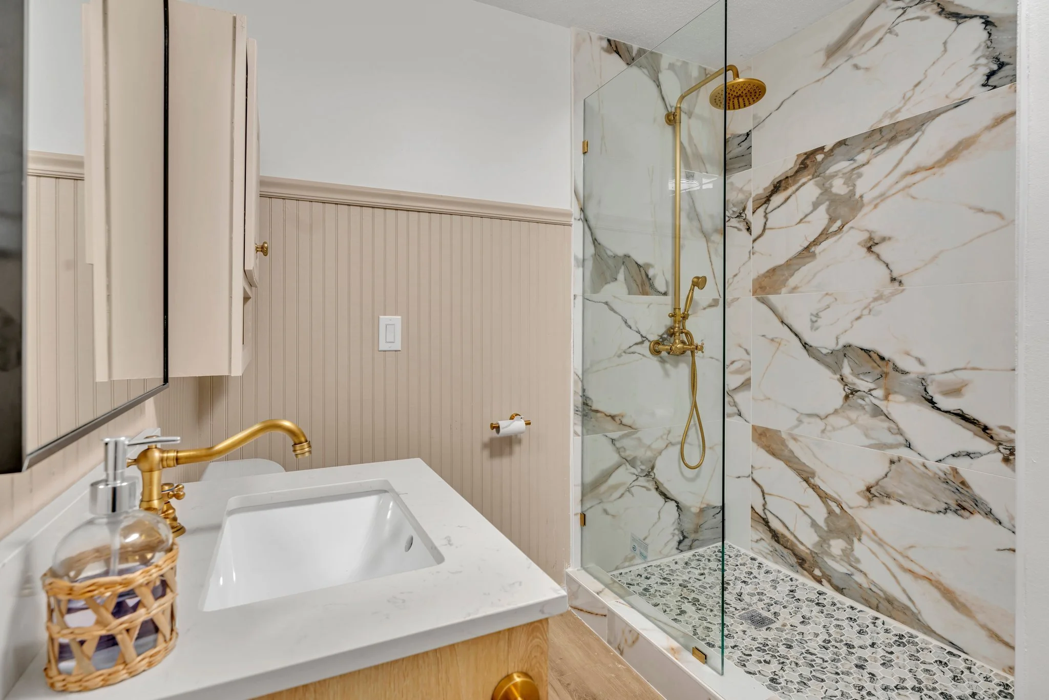Bathroom with beige beadboard walls, white countertop with gold faucet, small wicker basket, and a large shower with marble tiles, glass door, and gold fixtures.