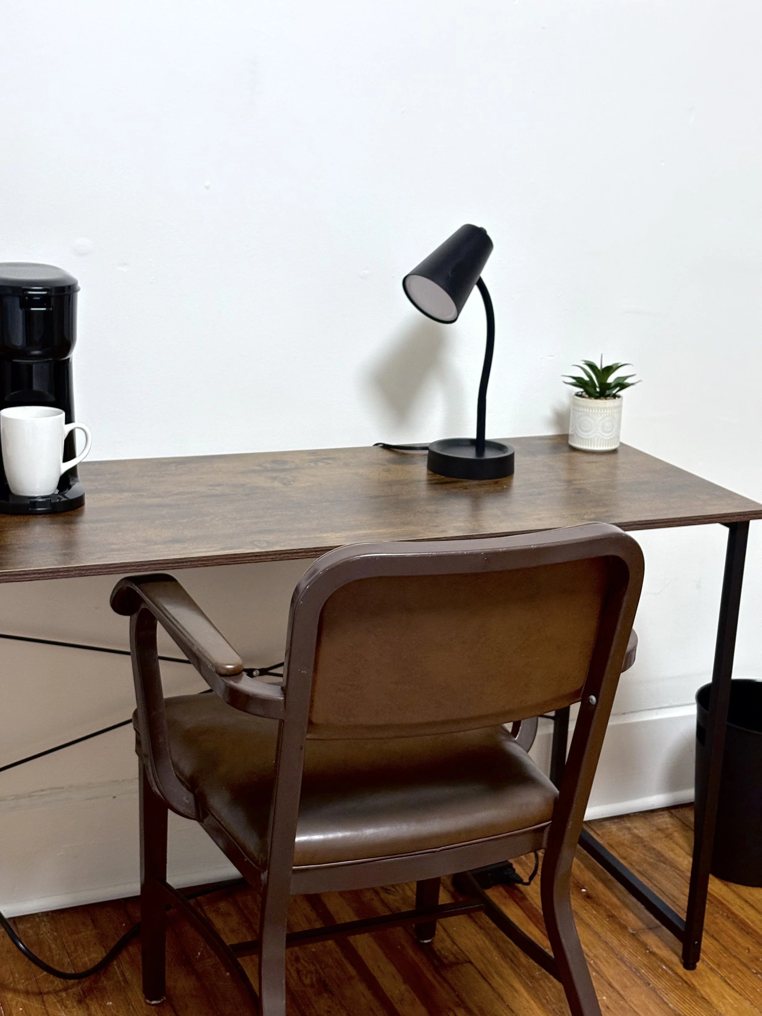 A wooden desk with a black desk lamp, a potted plant, a coffee maker with a white mug, and a black trash can underneath. A brown chair with a cushioned seat is in front of the desk. The wall behind the desk is white.