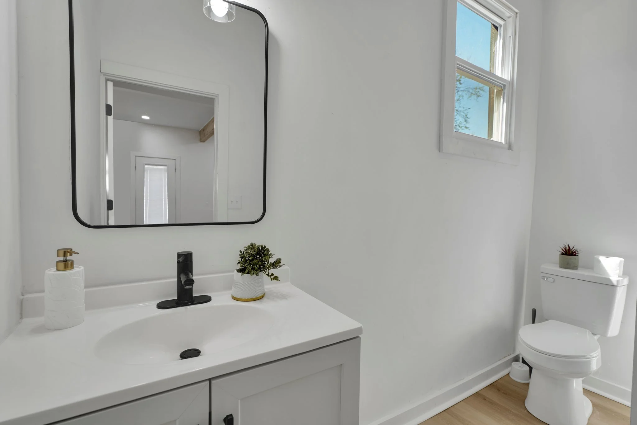 A minimalist bathroom with white walls, a white sink with black faucet, a mirror above the sink, a small potted plant, a soap dispenser, a window for natural light, and a white toilet with a small potted plant and toilet paper on top.