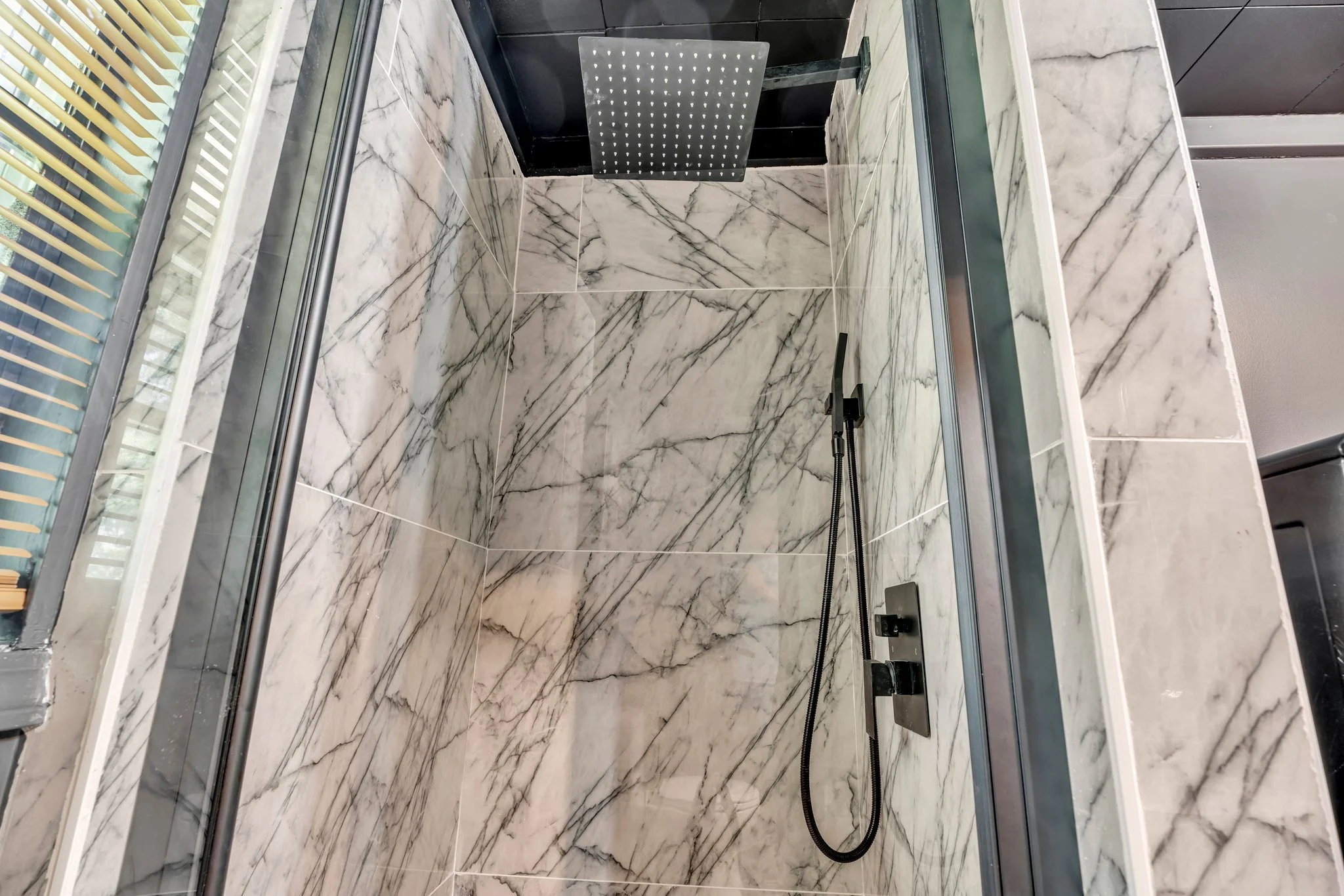 Marble tiled shower with a rainfall showerhead and a handheld shower attached to a black wall panel.