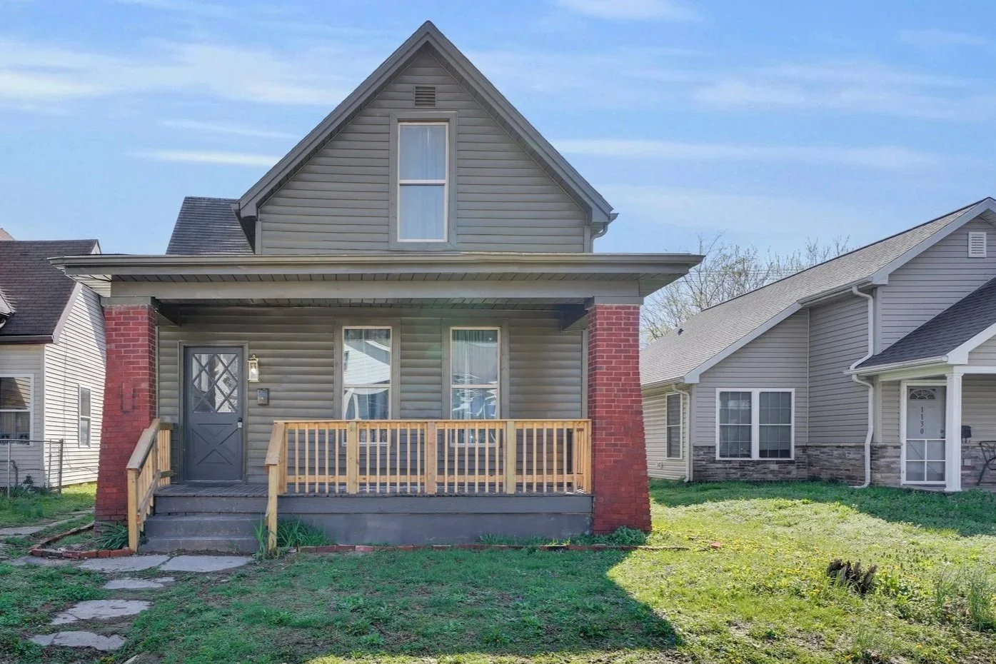 Front view of a gray two-story house with a wooden porch, brick pillars, and a small front yard.