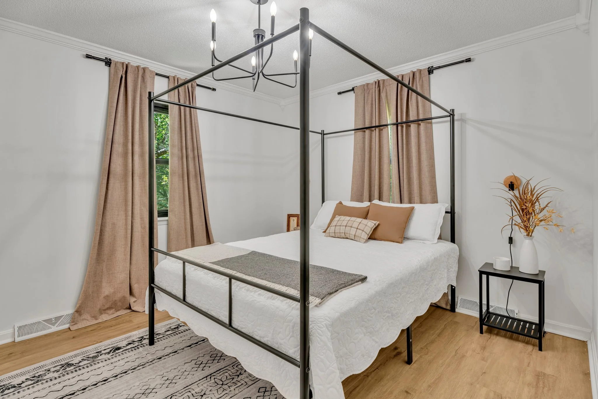 A bedroom with a metal four-poster bed, white bedding, beige pillows, and light brown curtains.