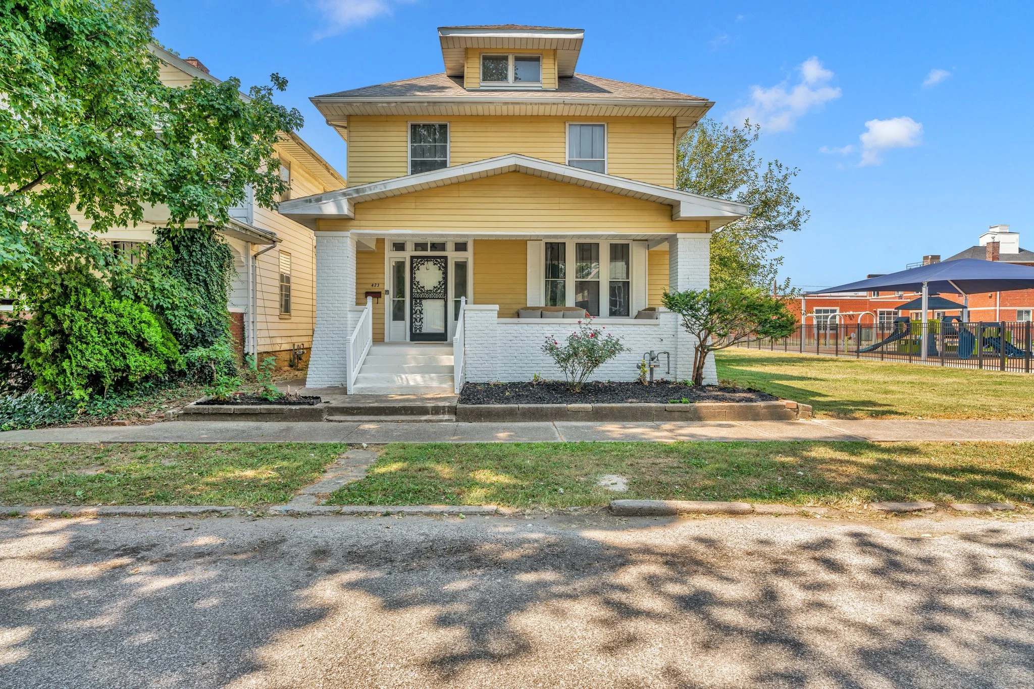 Front view of a yellow, three-story house with white trim and a front porch, surrounded by a yard with trees and a sidewalk, under a blue sky with a few clouds.
