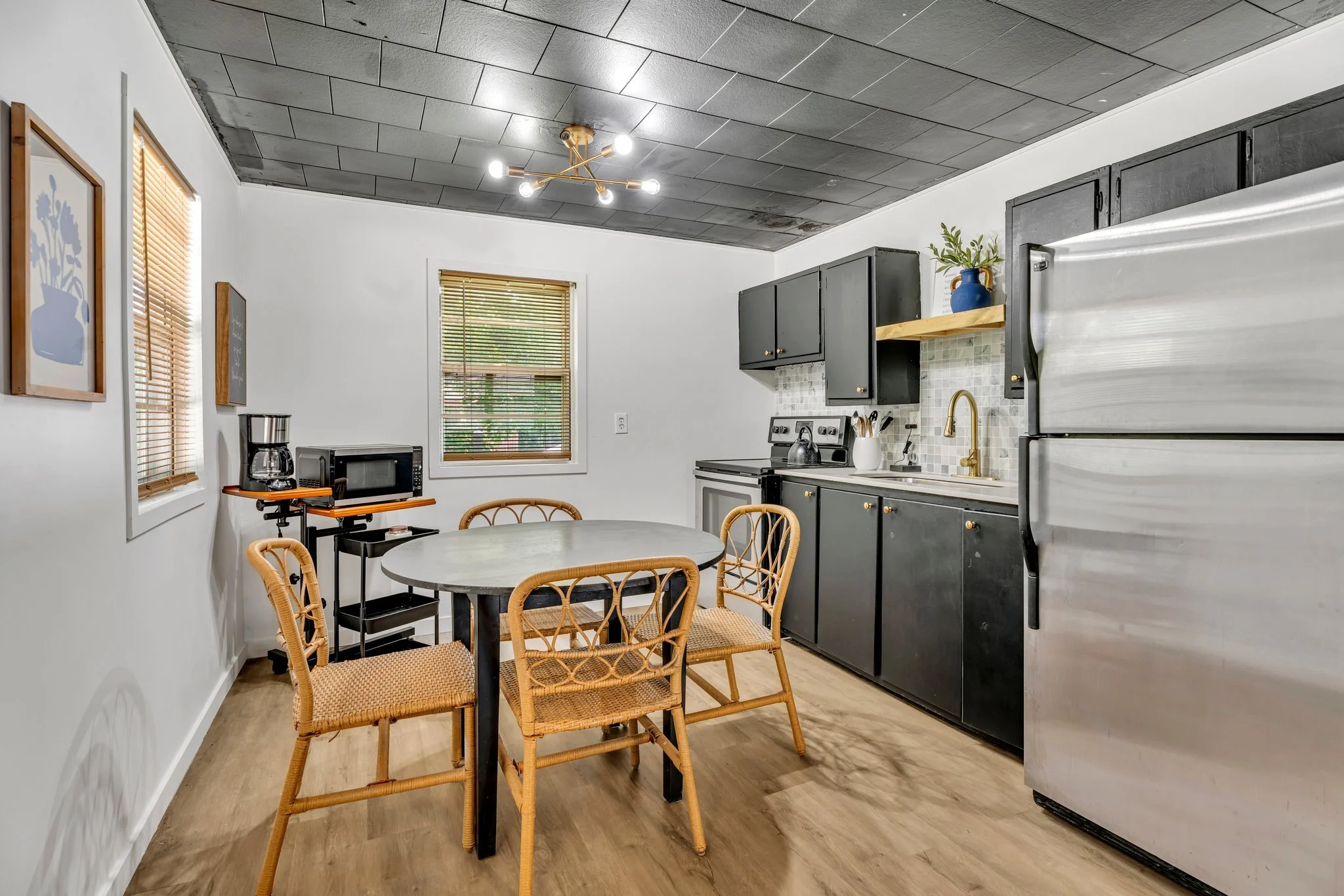 Kitchen with white walls, wooden flooring, black cabinets, stainless steel refrigerator, small round table with four wicker and rattan chairs, window blinds, microwave, coffee maker, blender, white sink with gold faucet, decorative blue vase with green plant, modern brass ceiling light fixture.